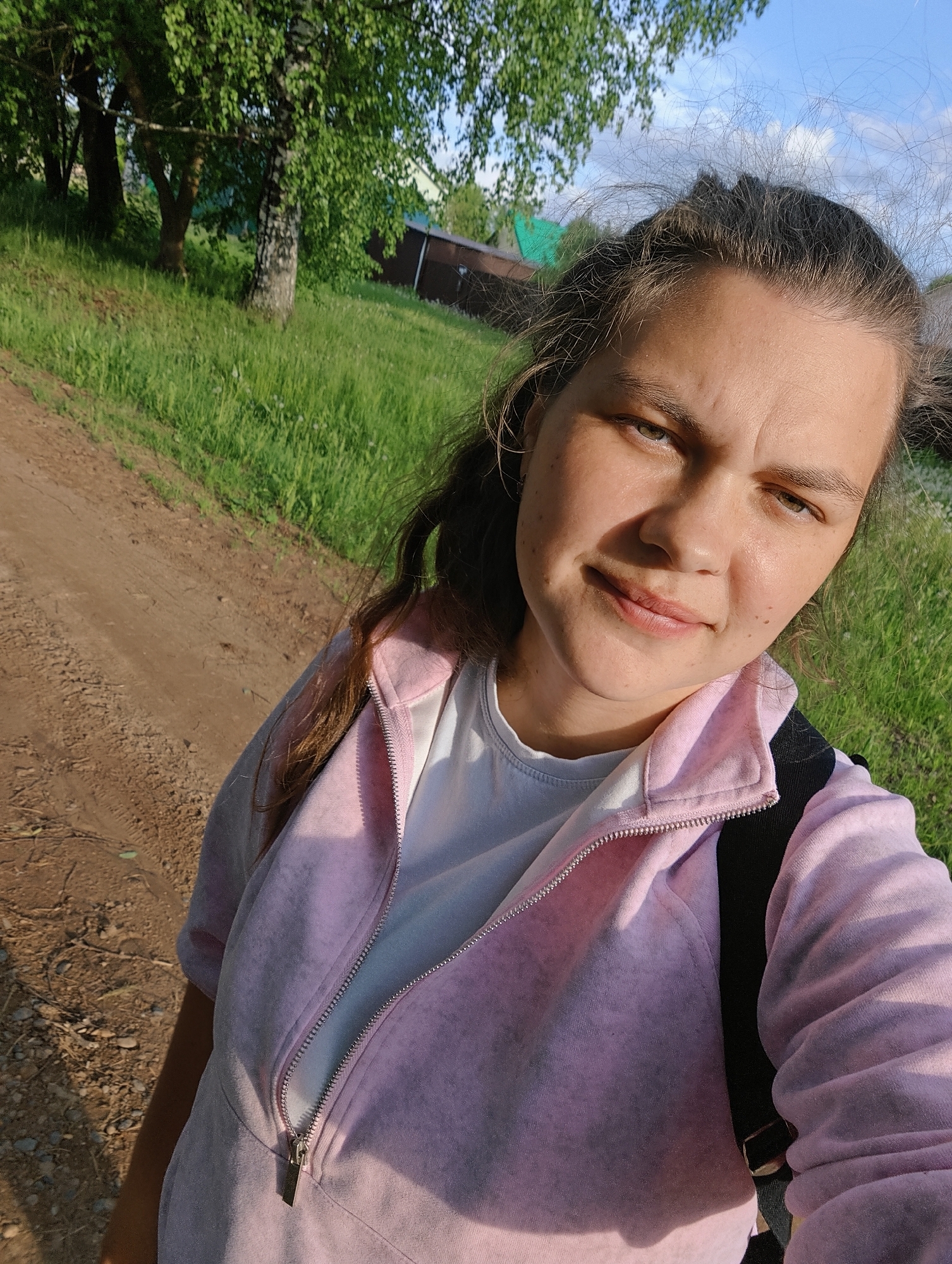 A medium, eye-level close-up shot depicts a woman in a casual pink zip-up jacket over a white t-shirt, with a black backpack strap visible on her shoulder. She is situated in what appears to be a rural or semi-rural setting on the outskirts of Уральское, Russia. A dirt path stretches beside a grassy area with trees in the background, indicative of a natural environment just outside urban development. A small, dark-colored building is partially visible behind the trees. The lighting suggests it is daytime. The woman is looking directly at the camera with a slight smile.