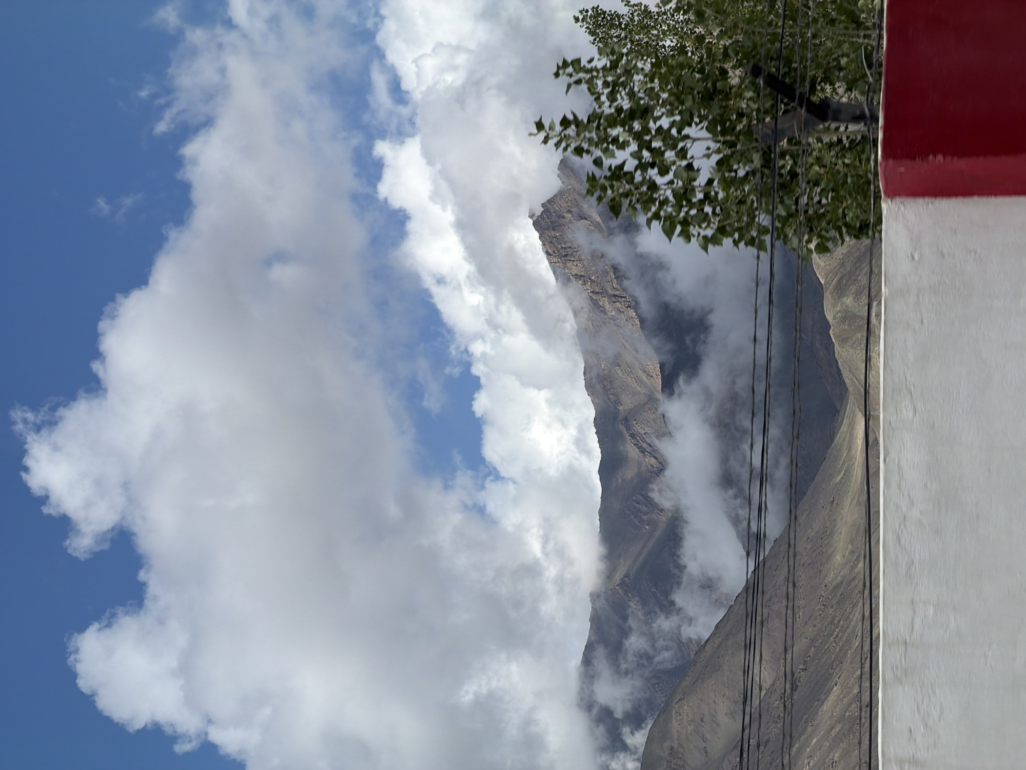 The image captures a mountainous landscape under a partially cloudy blue sky, near Spituk, India. The foreground features a concrete barrier, painted with white and red sections, and barbed wire fencing with a wooden post. Beyond the barrier, electrical wires are strung horizontally across the frame. The midground is dominated by the rugged slopes of mountains, partially obscured by drifting clouds that weave through the peaks and valleys. A tree with green leaves is visible on the right side of the image, adding a touch of vegetation to the scene. The overall impression is one of a remote, high-altitude environment, where natural elements meet man-made infrastructure.