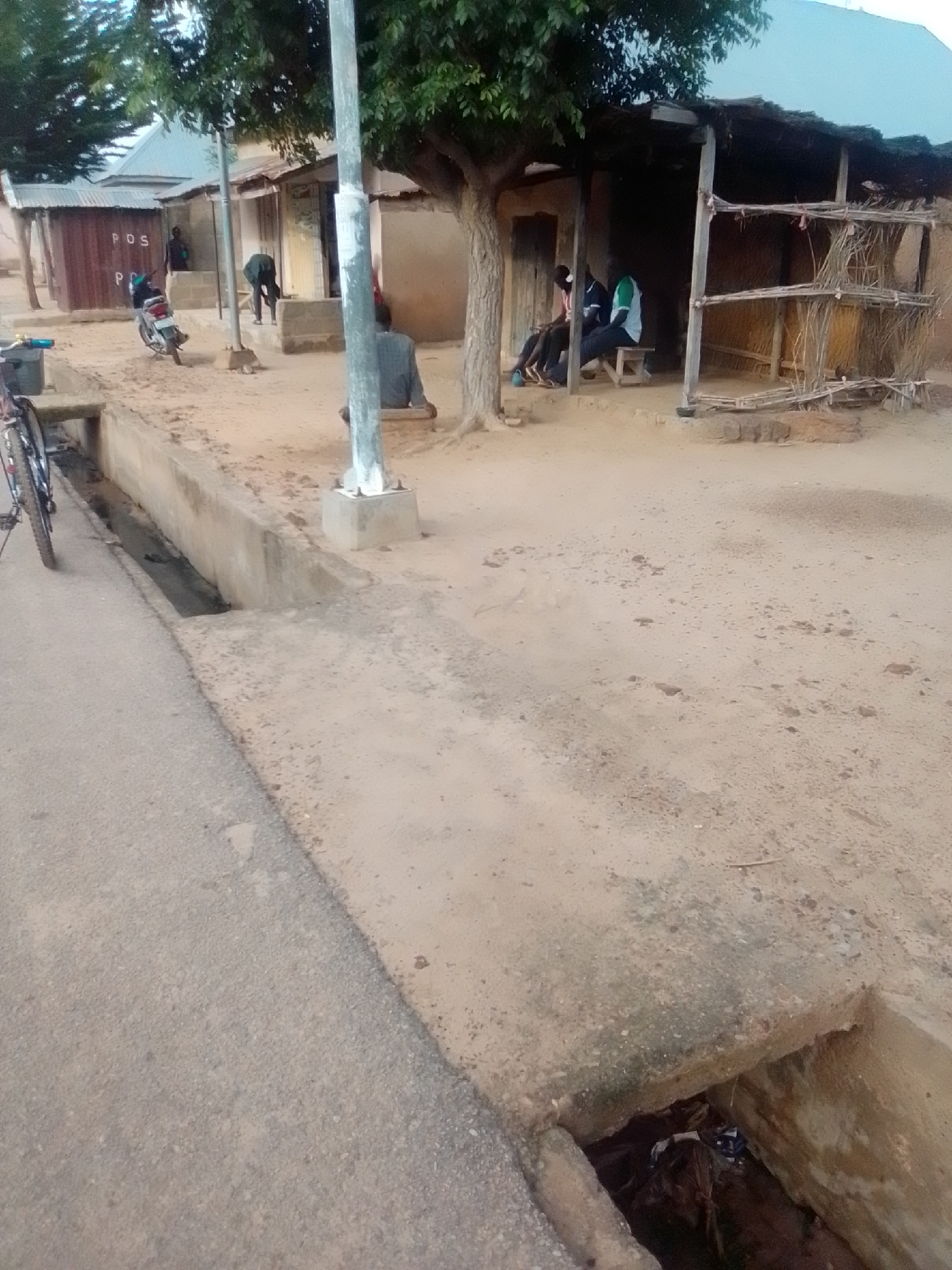 In the city of Gombe, Nigeria, an outdoor scene unfolds with a dusty, unpaved ground surface. A concrete drain runs along the left side of the image, with a bicycle parked next to it. In the background, several structures are visible, including a building with a corrugated metal roof and a sign that reads "P.O.S." Adjacent to this building is a more rustic structure with a thatched roof, held up by wooden poles. People are present in the scene: one individual is sitting on a stool in front of the P.O.S. building, another is bent over, and several others are seated under the thatched-roof structure. A motorcycle is parked in front of the P.O.S. building. The environment appears to be a street or thoroughfare within a residential or commercial area.