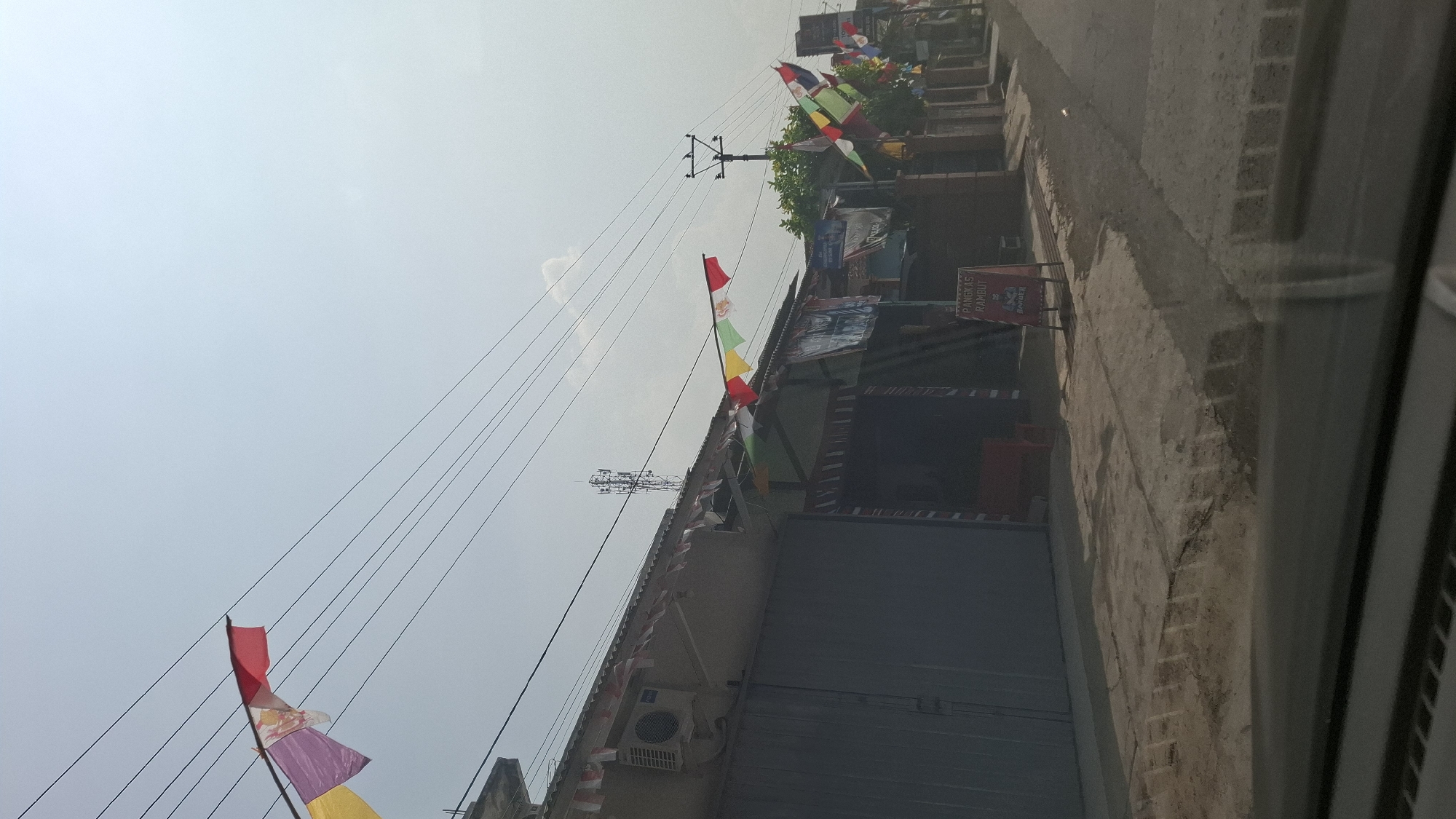 This is a low-angle, vertical, medium shot captured from inside a vehicle, looking out at a street scene in Sukasari, Indonesia. The sky is bright and hazy, with multiple power lines crisscrossing the frame. Along the rooftops of the buildings on the left, there are decorative flags strung across. The buildings appear to be small shops or residences, with corrugated metal roofs and dark-colored garage doors. An air conditioning unit is visible on the left building. A cell tower stands prominently in the background. Further down the street on the right, more colorful flags are displayed, and a barber shop sign is visible. The road surface is concrete and appears somewhat worn. The lighting suggests it is daytime, possibly mid-morning or afternoon, with the sun casting shadows on the ground. There are no people clearly visible in the image.