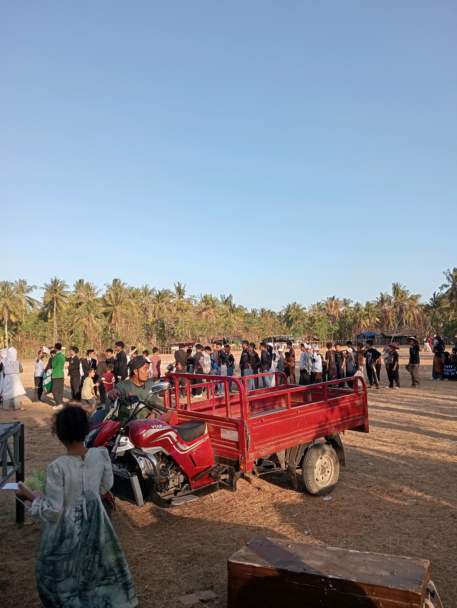 In Pagerungan Kecil, Indonesia, a group of people are gathered outdoors during daylight. The scene is set against a backdrop of palm trees and a clear blue sky. In the foreground, a young girl in a patterned dress walks away from the camera, holding some green items. To her right, a red three-wheeled vehicle is parked. The vehicle has a motorcycle-like front with a rider wearing a green shirt and helmet, and a flatbed cargo area at the rear. A crowd of people, predominantly young males dressed in a mix of casual and formal attire, are standing in groups to the left and in the background. Some individuals are wearing traditional Islamic clothing, such as caps and headscarves. The ground is covered in dry grass and dirt.