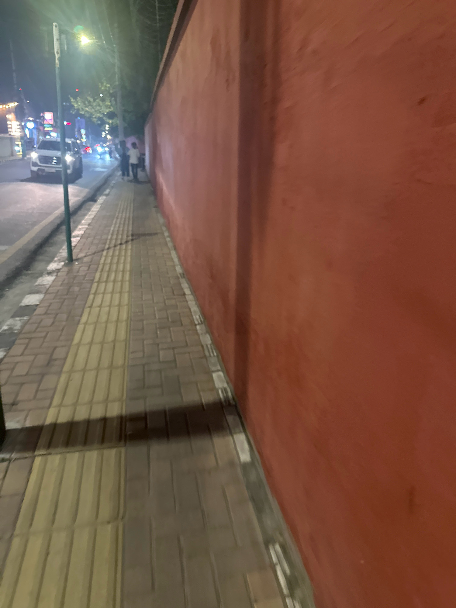 The image shows a sidewalk with textured tiles, possibly for the visually impaired, running alongside a solid, red-painted wall. The wall extends to the right of the frame and appears to be a boundary. The sidewalk is paved with rectangular bricks. To the left of the sidewalk, there is a street with vehicles and streetlights, indicating an urban environment at night. A white car is visible on the street. Several people are walking on the sidewalk and street. The overall scene suggests a street in Lalitpur, Nepal, at night, with a focus on the sidewalk and the adjacent wall. The lighting is artificial, coming from streetlights and vehicle headlights.