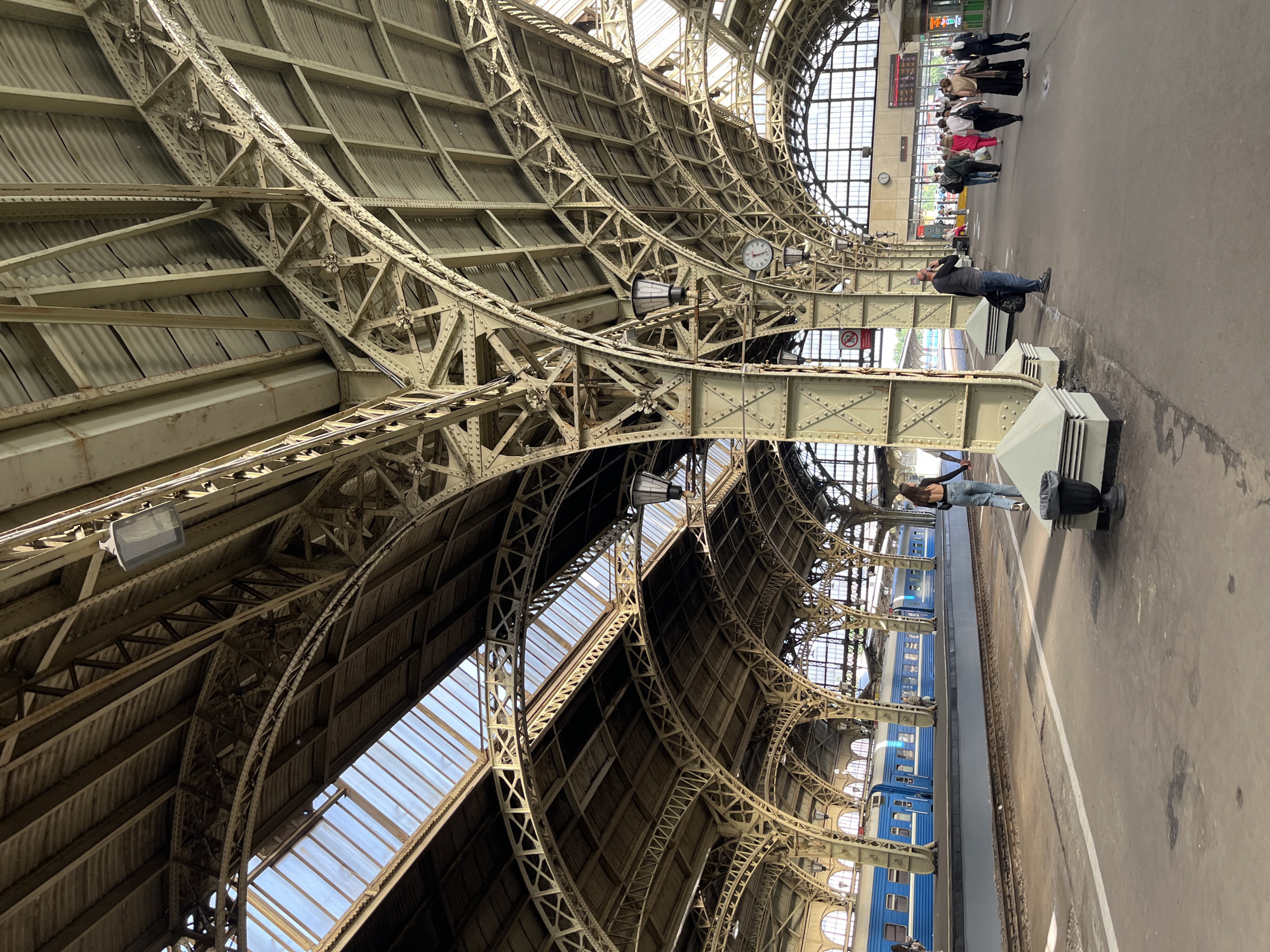 The image depicts the interior of a large, historic train station in Saint Petersburg, Russia, specifically Vitebsky Railway Station, which is known for its distinctive Art Nouveau architecture. The station's expansive roof is supported by an intricate network of yellow-painted metal beams and arches, with large glass panels allowing natural light to illuminate the space. Below, a train with blue carriages is visible on the tracks. Several people are present on the platform and concourse. In the foreground, a woman in jeans and a dark top walks away from the camera. Further back, a man in a dark shirt and jeans stands talking on his phone, holding a bag. A group of people, dressed in various casual and formal attire, walk further into the station. A large clock is mounted on a pillar. The overall impression is of a bustling, architectural landmark.
