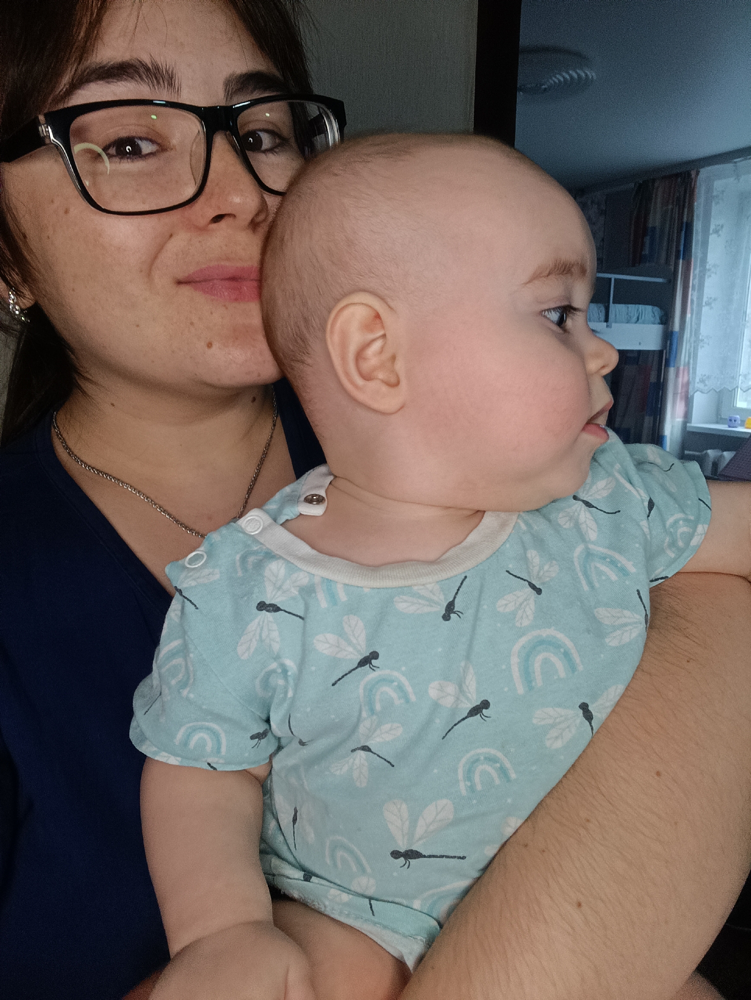 A woman with dark hair and glasses holds a baby in a light blue shirt with dragonfly and rainbow patterns. The woman wears a dark blue shirt and a silver chain necklace. The baby is looking to the right, and the woman is looking at the camera. The background includes a window with curtains and possibly a bunk bed. The location is Noviy, Russia.