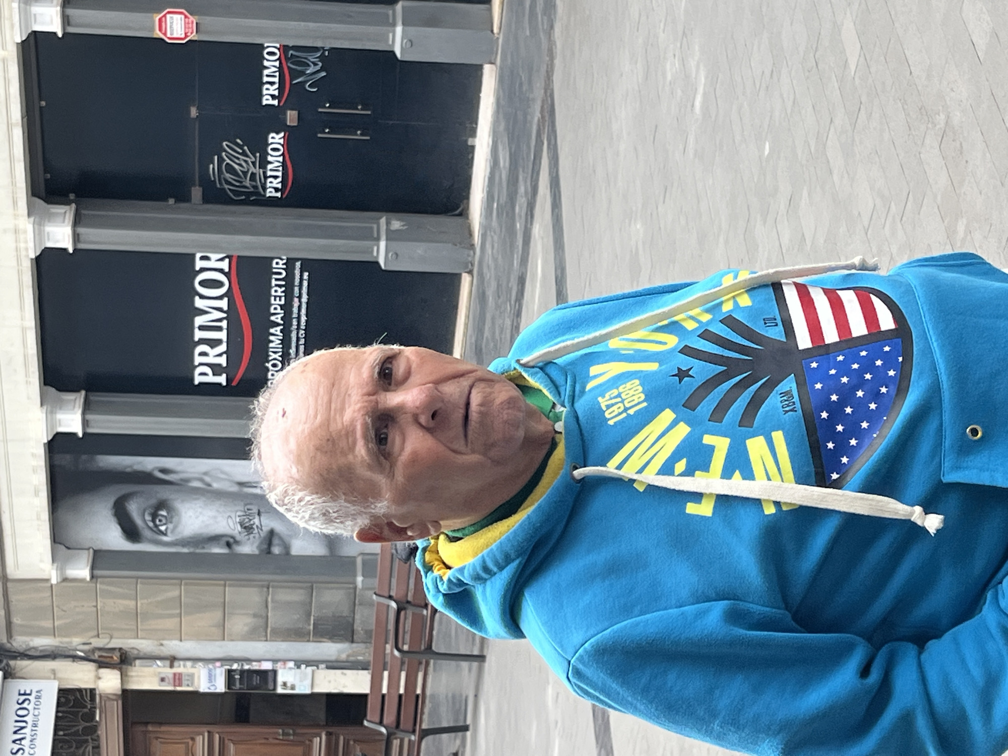 An elderly man with light skin and short, white hair is the central subject of the image, looking directly at the camera with a neutral, slightly curious expression. He is wearing a bright blue hooded sweatshirt with a yellow interior lining visible around the neck. The hoodie features a prominent graphic design on the chest, including elements reminiscent of the American flag (red and white stripes, blue field with white stars), alongside text such as "1975", "1986", and "XB&M". A white drawstring hangs from the hood.
The scene is set outdoors on what appears to be a paved street or pedestrian area in Las Palmas, Spain. The background is dominated by the dark facade of a commercial establishment. The name "PRIMOR" is prominently displayed multiple times on the storefront. Below one "PRIMOR" sign, Spanish text reads "PRÓXIMA APERTURA" (Next Opening), followed by recruitment details: "interesado en trabajar con nosotros envianos tu CV cvprimor@primor.es". A large, black and white artistic image of a person's eye and lower face, marked with some graphic elements, is visible on the store window to the man's left. A small red hexagonal sign is also affixed to the facade. Further to the left, a partial sign for "SANJOSE CONSTRUCTORA" is visible, and a wooden bench can be seen in the background. The scene is brightly lit by natural daylight, suggesting midday, with no strong shadows, indicating possibly an overcast sky.