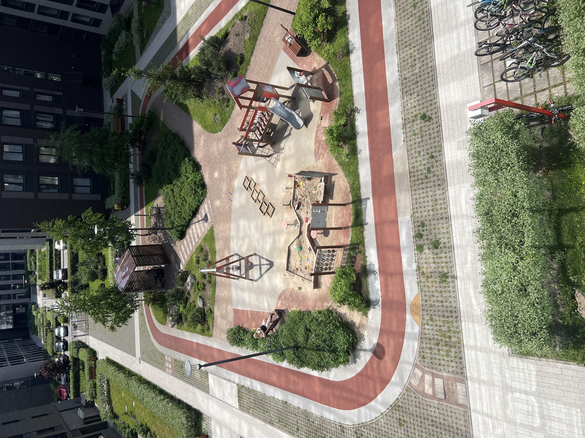 The image shows a modern residential courtyard in Saint Petersburg, Russia, featuring a playground, walking paths, green spaces, and parking areas. The courtyard is designed with a combination of paved surfaces and landscaped areas. A playground with a slide, sandbox, and climbing structures is centrally located. Walking paths, some with red surfacing, wind through the courtyard. Greenery, including bushes and trees, is abundant, and there are several benches for seating. In the background, modern apartment buildings are visible, as well as parked cars and a bicycle rack with multiple bicycles. The scene appears to be an outdoor urban recreational space.
