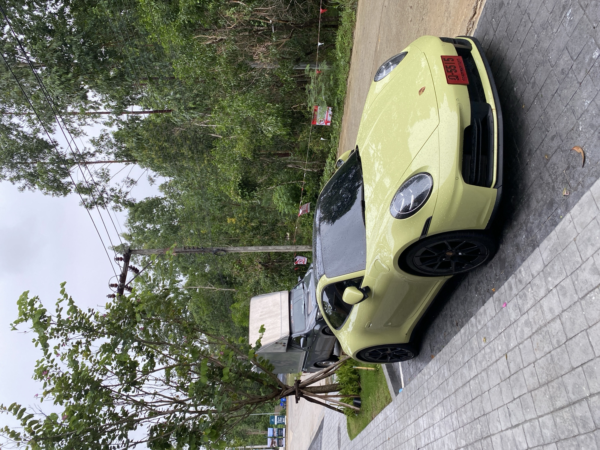 A lime green Porsche coupe is parked on a concrete paver surface adjacent to a lush, tropical green space with trees and foliage. A utility pole with power lines is visible against a grey sky. A silver pickup truck with a white box enclosure is parked behind the Porsche. The license plate on the Porsche reads "อ-5515," indicating the image location is likely Choeng Thale, Thailand. Small flags and a sign attached to a rope appear along the edge of the road near the Porsche. The car is wet, suggesting it has recently rained.