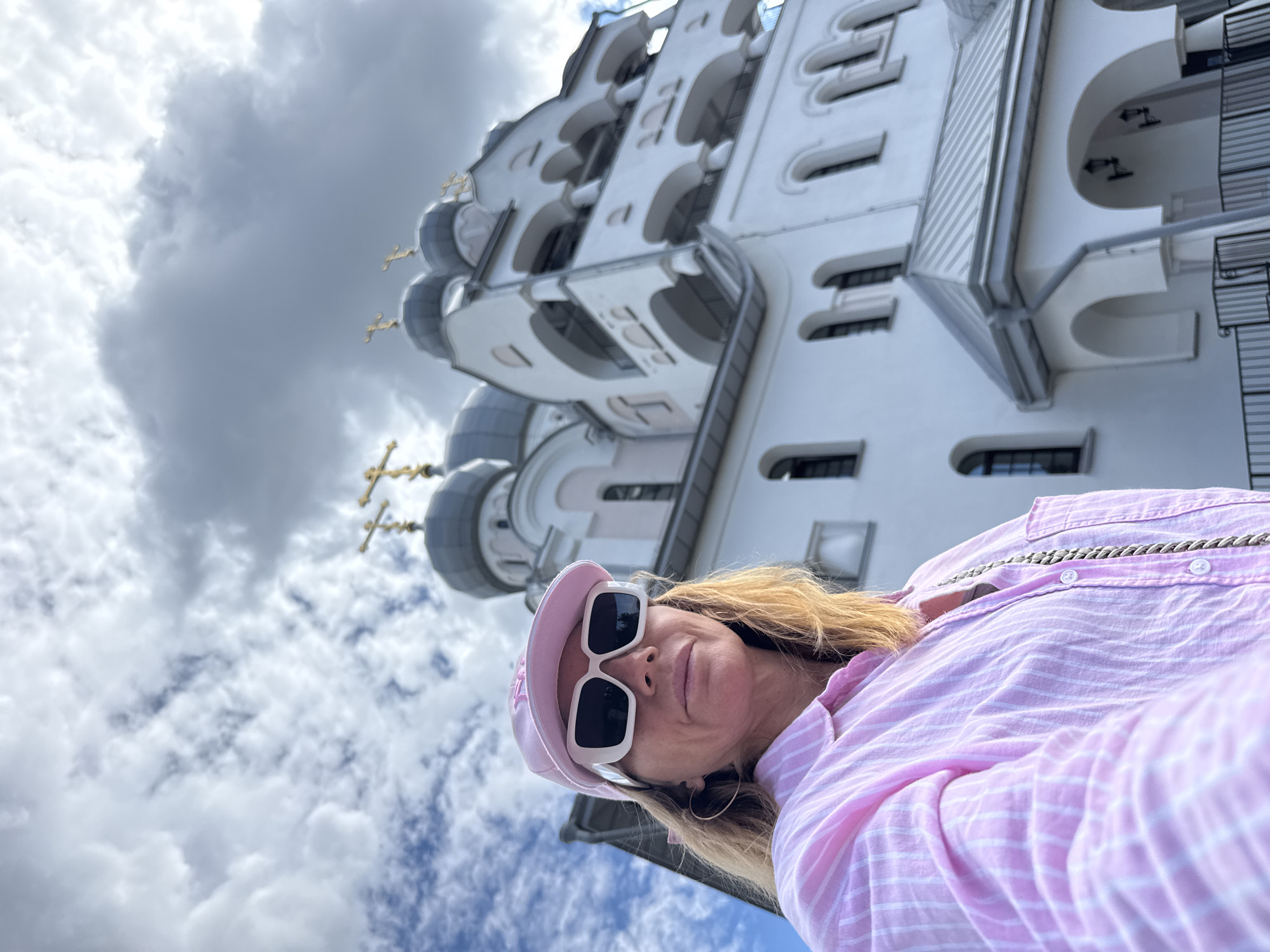 A woman wearing a pink striped shirt, a pink baseball cap, and white sunglasses takes a selfie in front of a large white church with gray domes topped with golden crosses. The church has arched windows and balconies. The sky is cloudy. The scene appears to be in Saint Petersburg, Russia, possibly near the Church of the Nativity of the Theotokos.