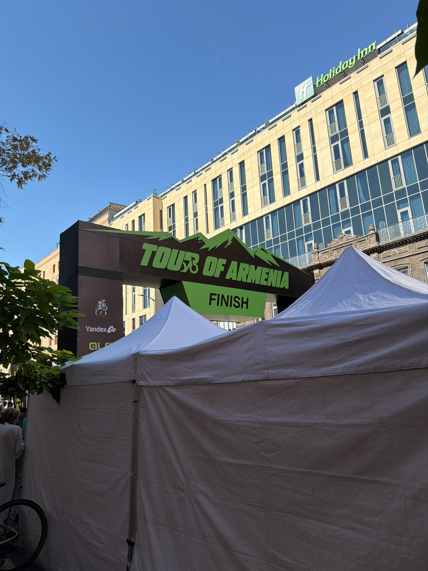 The image captures a sunny day in Yerevan, Armenia, with the "Tour of Armenia" finish line arch prominently displayed. The arch is a dark brown structure with green lettering that reads "TOUR OF ARMENIA" and "FINISH," framed by mountain graphics. Below the arch, white tents are set up, suggesting an event or race. In the background, the modern Holiday Inn hotel stands tall, its yellow facade and blue-tinted windows contrasting with the blue sky. To the left of the arch, a bicycle is partially visible, leaning against a white tent. Some people are seen in the background, near the entrance to the event area, but their faces are not clearly discernible. Trees with green leaves are also present, adding a touch of nature to the urban setting. The scene appears to be a street or plaza in Yerevan, likely a popular area for events and tourism.