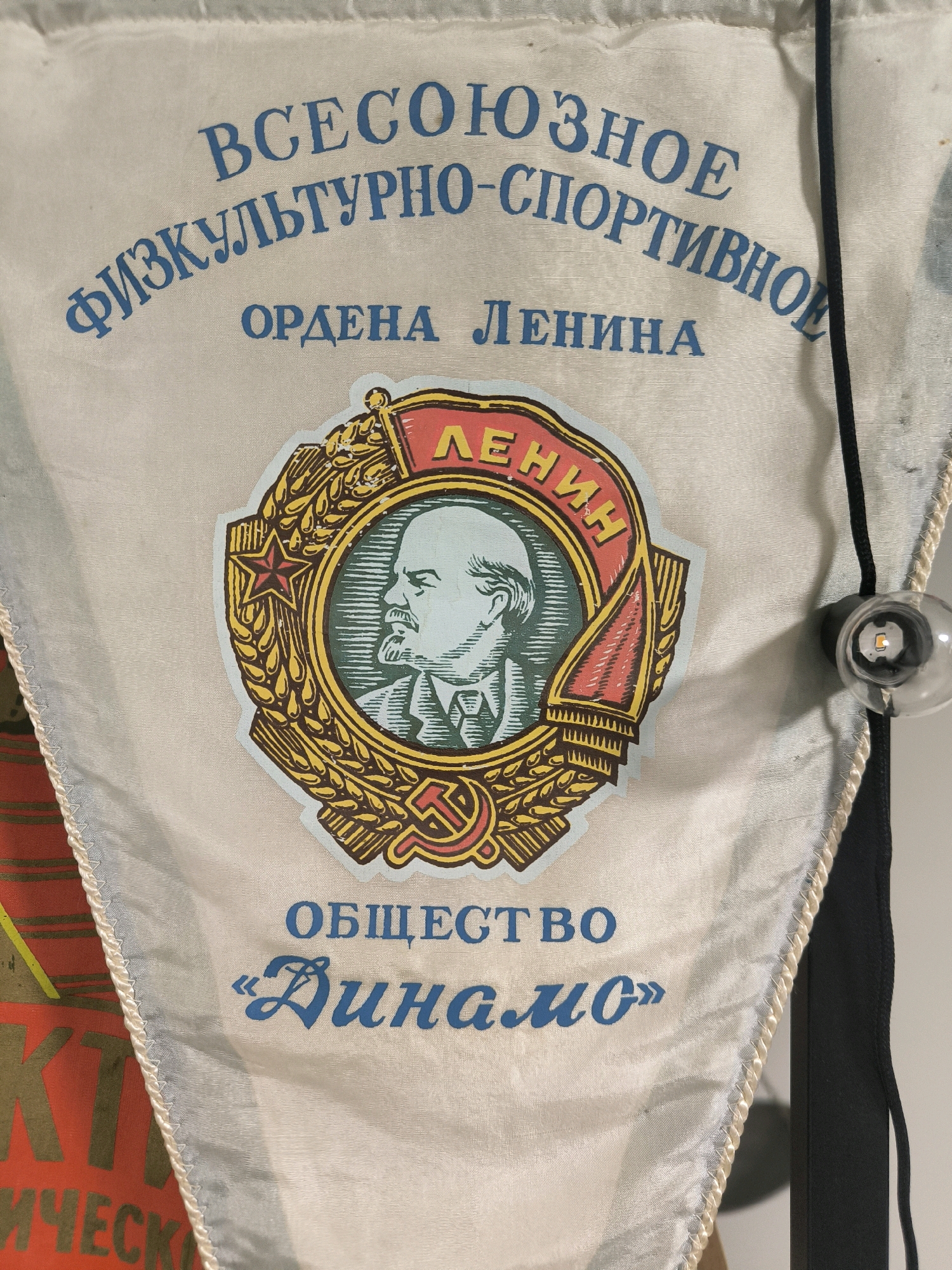 The image is a close-up of a white pennant with blue Cyrillic text and a circular emblem. The pennant appears to be made of fabric with a satin-like sheen. The text at the top, curved to follow the shape of the pennant, reads "ВСЕСОЮЗНОЕ ФИЗКУЛЬТУРНО-СПОРТИВНОЕ". Below this, also in blue, is "ОРДЕНА ЛЕНИНА".
Centered on the pennant is a circular emblem depicting a profile of Vladimir Lenin, rendered in light blue and green tones. He is depicted with a beard and mustache, wearing a suit. The emblem is surrounded by a wreath of golden wheat stalks and a red banner that reads "ЛЕНИН" in yellow. At the bottom of the emblem, a red hammer and sickle symbol is visible.
Below the emblem, the blue text reads "ОБЩЕСТВО" and then, in a more stylized, script-like font, "«Динамо»".
The pennant has decorative braided trim along its edges. A black cord runs vertically along the right side of the pennant, and a small, clear light bulb with an LED element inside is attached to this cord.
The background of the image is a plain, light-colored surface, likely a wall or a display. To the left of the pennant, a portion of another object is visible, appearing to be a red surface with large, yellow, blocky Cyrillic letters.
The image does not contain any information or visual cues that would allow for the determination of its location within the city of Magnitogorsk, Russia, or any specific environment. The scene is focused solely on the pennant and its details.