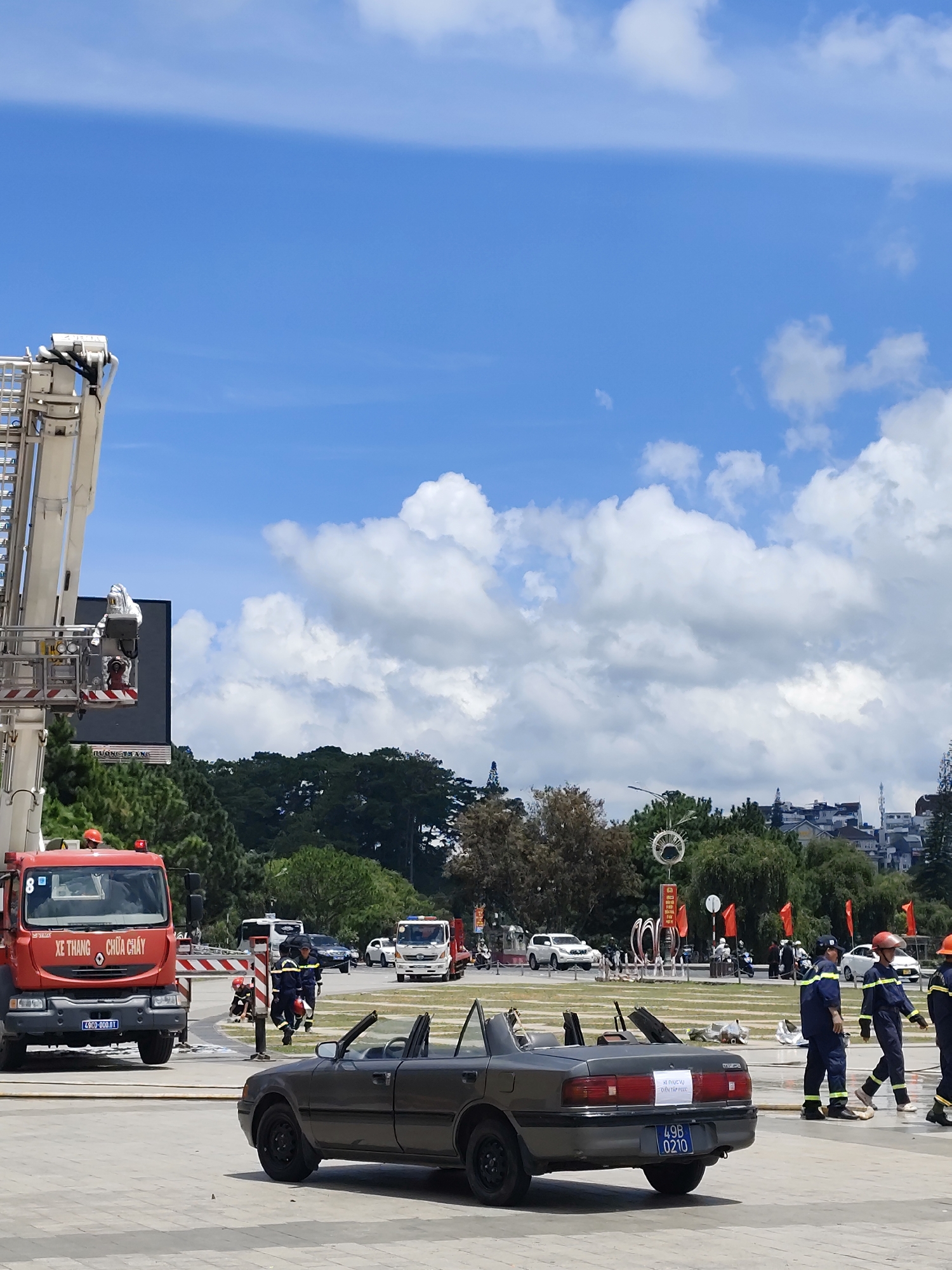 In Da Lat, Vietnam, a scene unfolds involving emergency personnel and vehicles. A large ladder truck, painted bright red, is positioned on the left, its ladder extended upwards and a person in a silver protective suit visible in the basket. Adjacent to the truck, a digital billboard displays Vietnamese text, likely related to the event.
In the foreground, a grey convertible with its top down is parked, a white sign with Vietnamese text visible on its rear. Several individuals in blue and yellow emergency uniforms, some wearing red helmets, are present near the vehicles, appearing to be part of an operation or drill. Another red fire truck is visible further in the background, alongside other vehicles and a crowd of onlookers.
The environment suggests a public space, possibly a plaza or intersection, with trees and buildings in the distance. The sky is bright blue with scattered white clouds, indicating fair weather. The overall atmosphere suggests a public safety demonstration or response.
