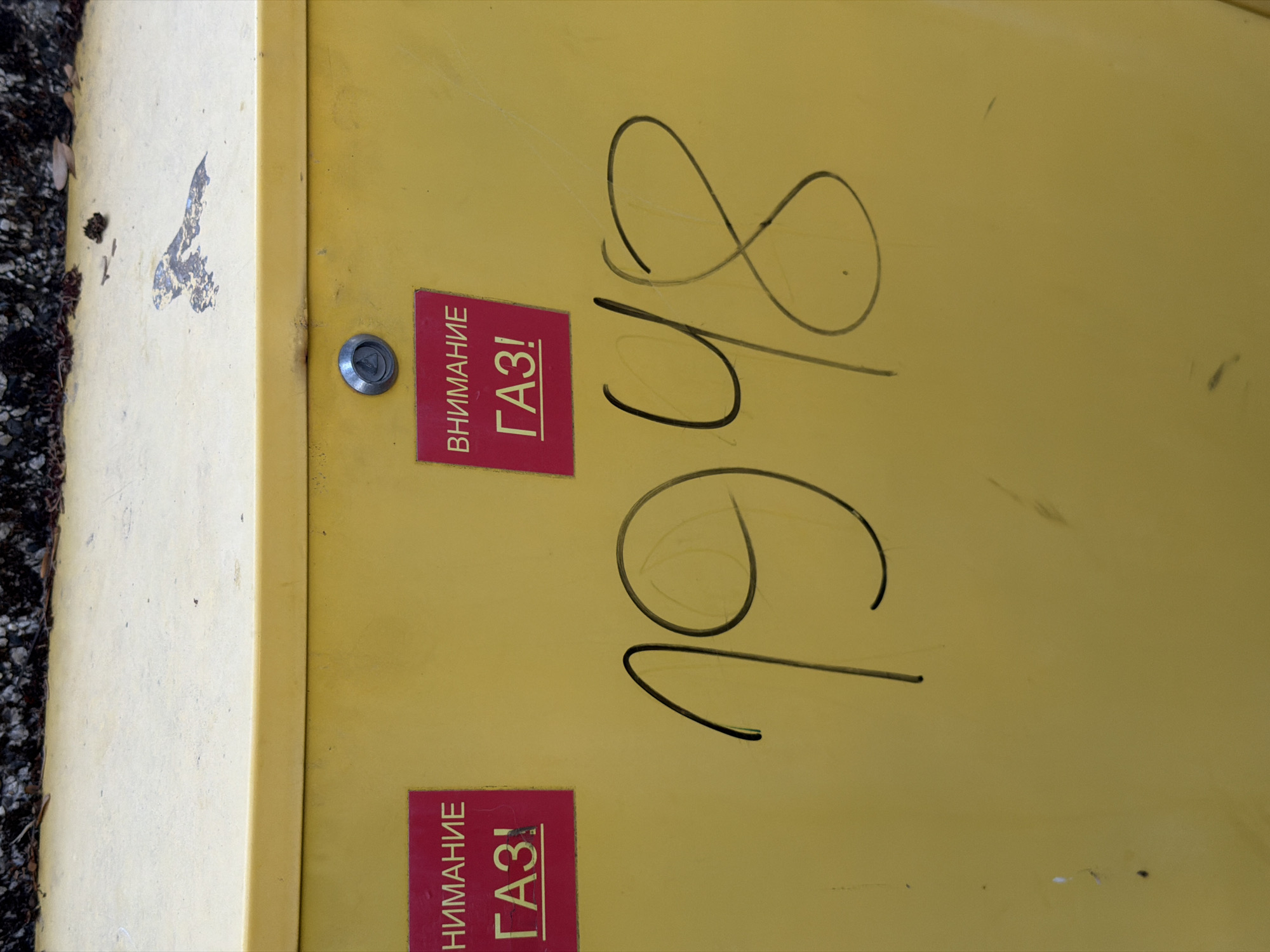The image shows a close-up of a yellow metal structure, likely a utility box, in Svilengrad, Bulgaria. The structure has two red warning stickers with Cyrillic text. The left sticker reads "НИМАНИЕ" (Attention) above "ГАЗ!" (GAS!). The right sticker has the same text. Large, hand-drawn numbers "1948" are scrawled in black marker across the front of the yellow surface, obscuring a portion of the lower part of the stickers. The top of the structure is visible, covered in a light-colored material, with some debris and a few dark specks. A metal lock mechanism is visible on the front of the yellow surface, slightly above and between the two stickers. The overall scene suggests an outdoor urban environment, though no specific landmarks or background elements confirm the exact location within Svilengrad.