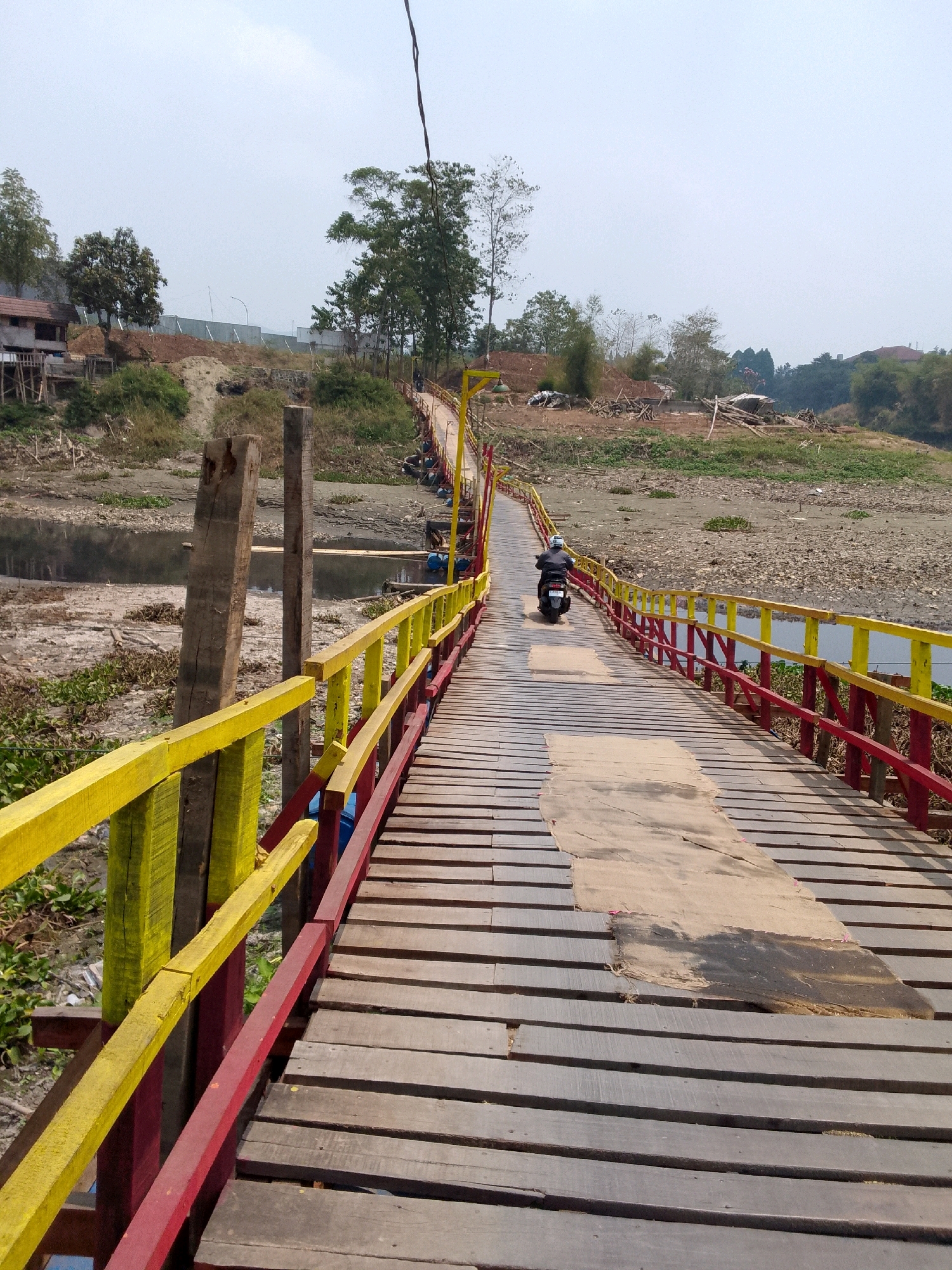 In Cipeundeuy, Indonesia, a weathered wooden bridge spans a dry riverbed. The bridge features bright yellow and red railings, contrasting with the aged, graying planks of the walkway. A patch of tan fabric is laid down on the bridge, appearing to cover damaged sections of the wood. A lone individual rides a motorcycle across the bridge, moving away from the camera. The surrounding environment is arid, with sparse vegetation and dry, cracked earth visible along the riverbanks. Hills and trees form the backdrop under an overcast sky.
