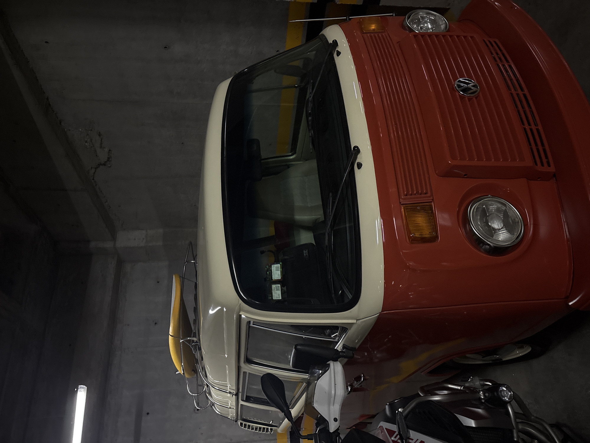 The image shows a red and white Volkswagen van parked in a parking garage in Jesús del Monte, Mexico. The van has a chrome VW emblem on the hood and a yellow surfboard strapped to the roof rack. A motorcycle is parked next to the van. The parking garage has concrete walls and yellow stripes on the columns.