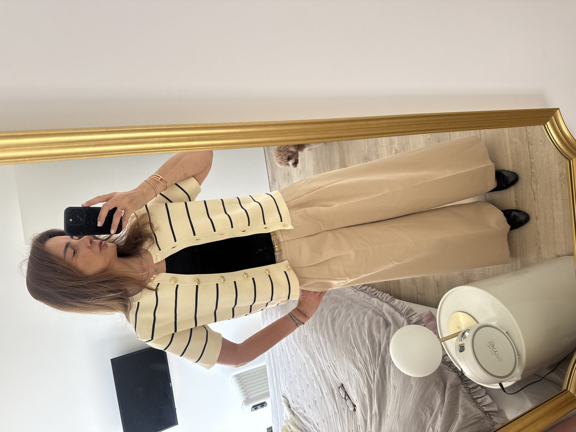 A woman is taking a photo of herself in a full-length, gold-framed mirror. She's wearing a striped cardigan, a black top, and wide-leg beige pants. She's holding a phone in her right hand and posing with her left hand on her hip. The mirror reflects a bedroom with a bed, a lamp, and a television on the wall. A dog is visible in the background. The location appears to be an interior space, potentially a home or apartment. It is not possible to determine if this space is in Vila Nova de Gaia, Portugal, without external information or visual landmarks indicating the location.