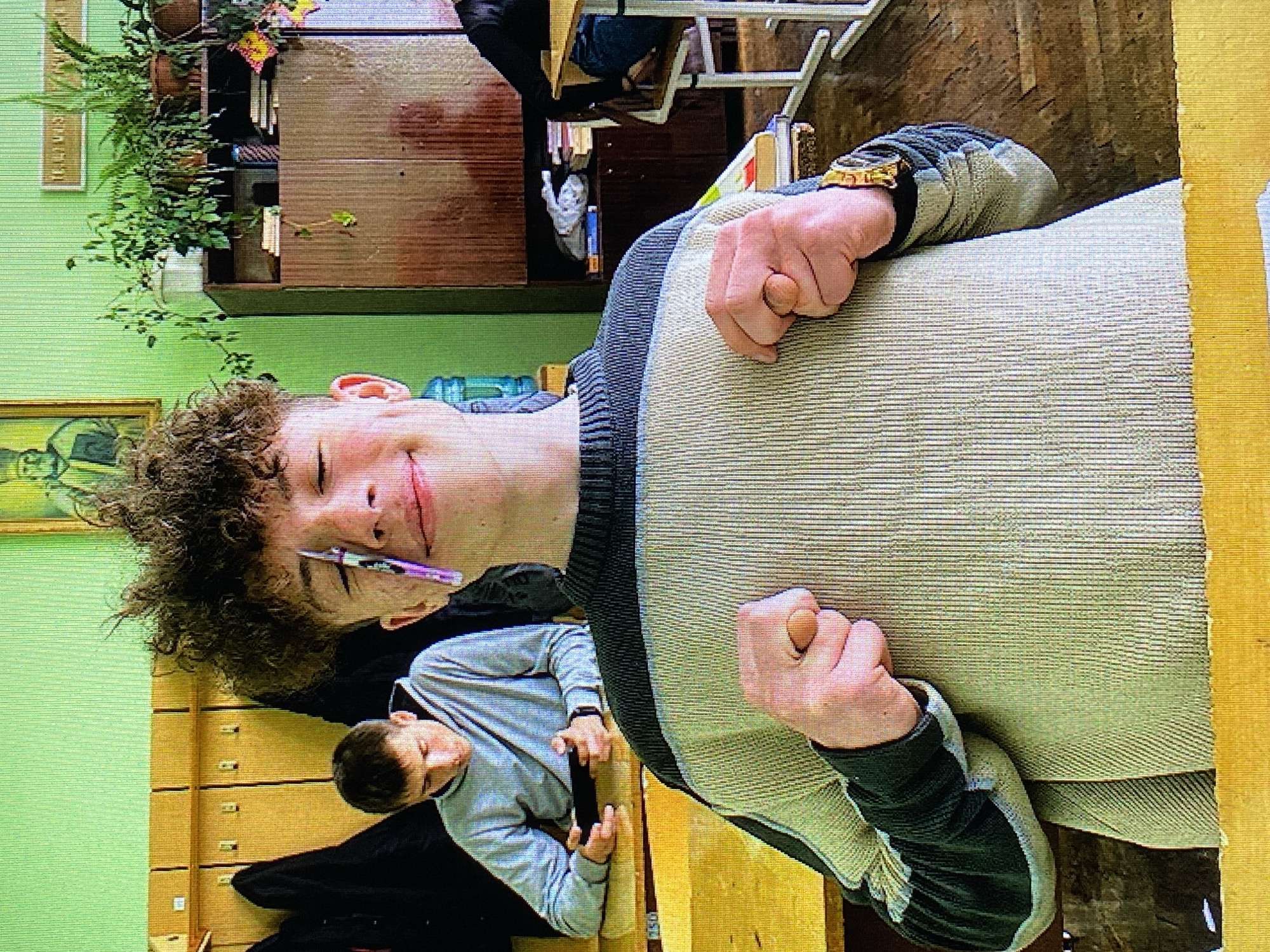 The image captures a young male with curly hair, wearing a striped sweater, positioned at a desk in what appears to be a classroom. He is holding his hands up in front of him, with his fists clenched. A pen is placed across his forehead, resting on his eyebrows. To his left, another young male in a grey hoodie is seated at a desk, looking down at a mobile phone. The room is painted green, with wooden lockers on the left wall and a wooden cabinet filled with plants and books on the right. The desks are made of wood with metal legs. The lighting suggests an indoor environment, possibly during daytime. The overall impression is that of a candid shot within a school setting. The specific location in Poznan, Poland, cannot be determined from the visual information provided.