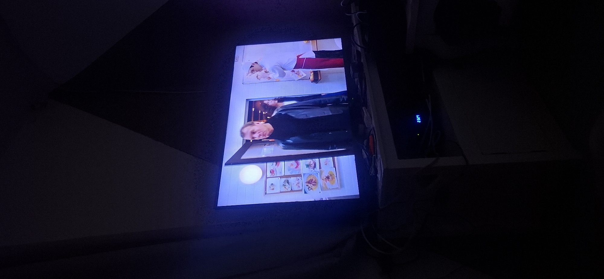The image shows a television screen displaying a scene with at least three people. A man in a dark turtleneck and a dark jacket is in the foreground, looking to his left. Behind him, a woman with dark hair is partially visible. To the right, a person wearing a chef's hat and a white coat with a red apron is visible. The background on the screen appears to be an interior scene, possibly a restaurant or kitchen, with framed pictures on the wall. The television is placed on a stand or table, with various electronic devices and cables visible below it. A digital clock on one of the devices shows "23:29". The overall environment is dark, suggesting the image was taken in a dimly lit room. This image is located in the city of Yerocham, Israel.