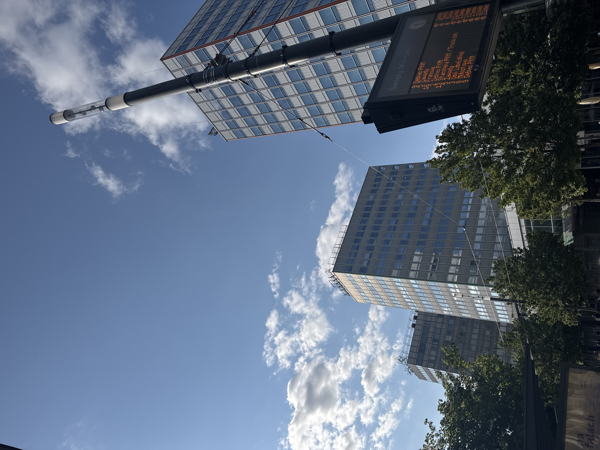 In Poznań, Poland, on a clear day with scattered clouds, a low-angle shot captures the modern architecture of the city. The focal point is a tall, dark pole with a cylindrical light fixture at its apex, extending upwards against the bright blue sky. Attached to this pole is a digital display board showing route information for "Święty Marcin" in Polish. Several glass-faced skyscrapers with grid-like window patterns dominate the background, some with reddish accents on their facades. Trees with lush green foliage are visible in the foreground, partially obscuring the base of the buildings. The scene suggests a public transport stop or a civic space within a contemporary urban environment.