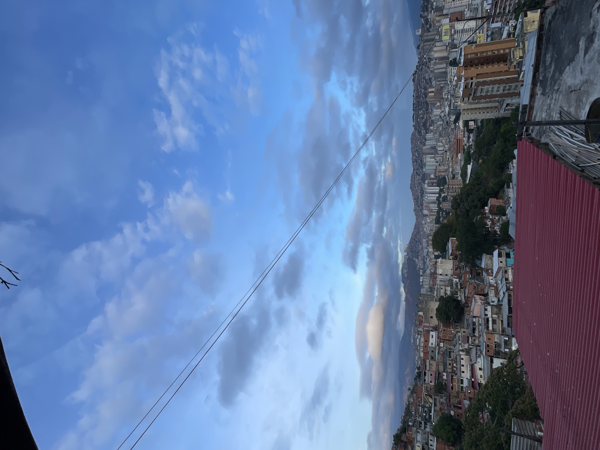 The image presents a view in Caracas, Venezuela. The scene is divided between a cloudy blue sky and a cityscape. The cityscape shows a mix of densely packed low-rise structures and taller buildings extending toward a mountain range in the background. A red corrugated metal roof and a metal railing are visible in the foreground. Power lines are present in the upper portion of the image. The overall environment suggests an elevated vantage point overlooking a densely populated urban area.