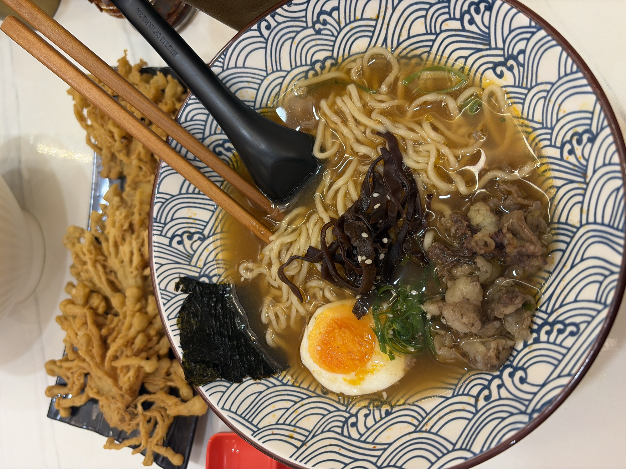 A close-up, eye-level shot captures a bowl of ramen with various toppings, accompanied by a side dish of fried enoki mushrooms. The ramen, served in a decorative blue and white patterned bowl, features noodles, a soft-boiled egg with a vibrant orange yolk, pieces of beef, green onions, and dark seaweed. Chopsticks rest on the rim of the bowl, with a black ladle partially submerged in the broth. The fried mushrooms are presented on a black rectangular plate in the background. The overall setting appears to be a restaurant. There is no indication of the specific location within Pekalongan, Indonesia.