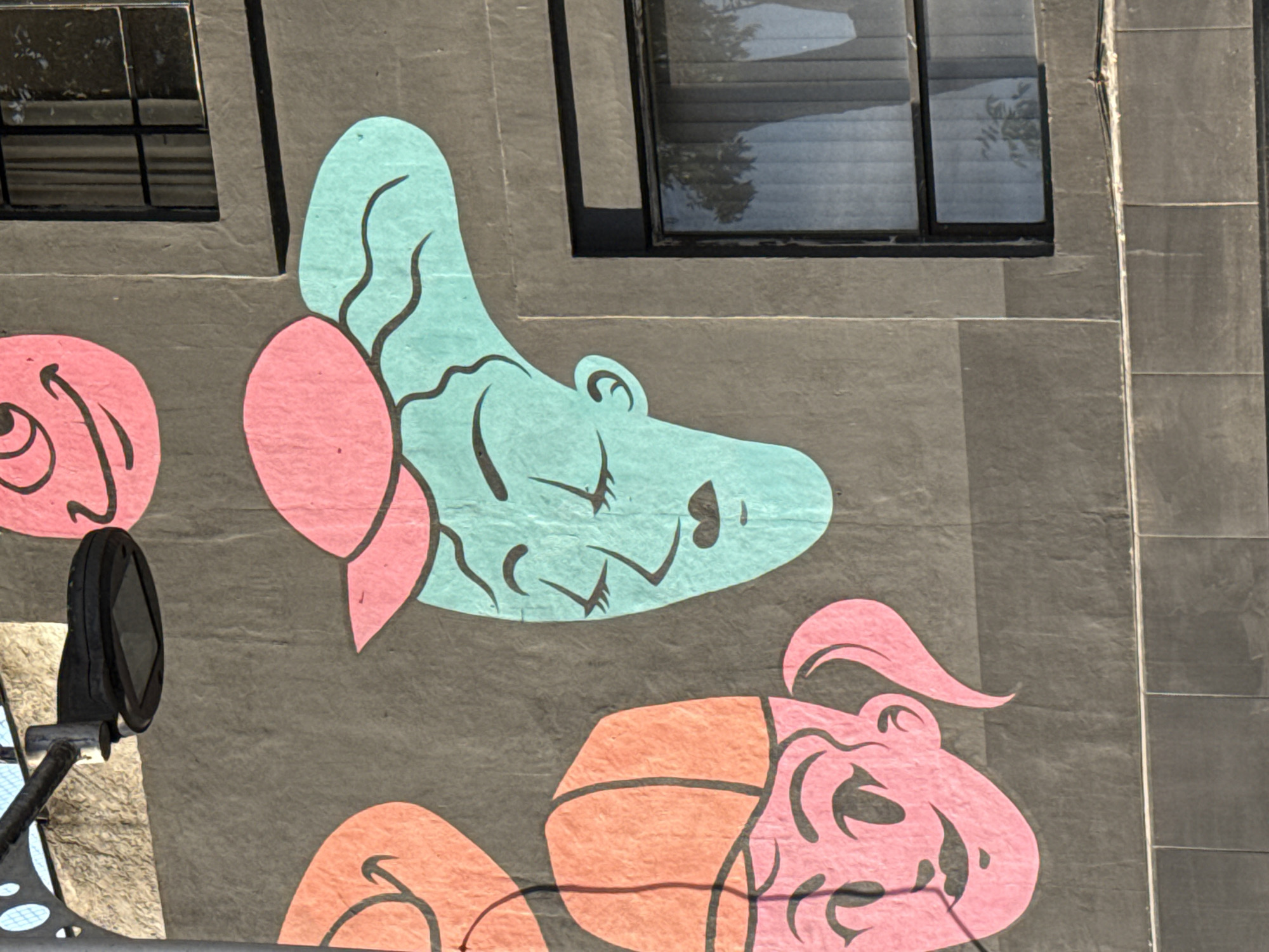 A portion of a mural on a building in the city of Puebla, Mexico, is seen. The mural consists of cartoonish renderings of hippopotamus-like figures in pink, orange, and blue colors against a grey background. The hippos are arranged vertically on the wall. Two black framed windows are positioned near the top of the image. A street lamp is seen in the bottom left of the image. The building appears to be constructed with textured grey stone or masonry.