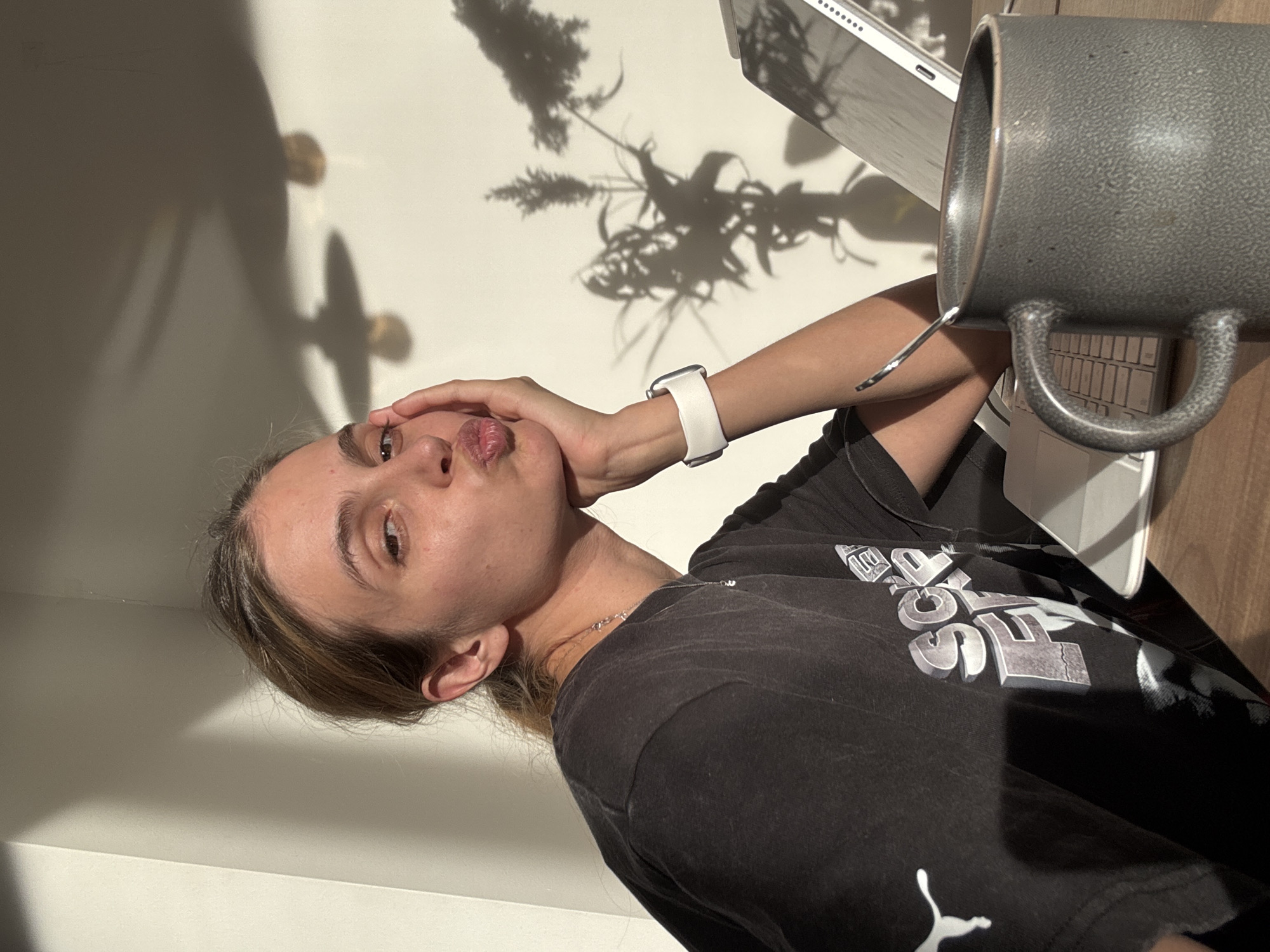A woman is posing with her hand on her cheek, pouting. She is wearing a black t-shirt, a white smartwatch, and a silver necklace. A large gray mug is to her right, and a laptop is behind the mug. Shadows of foliage are visible on the wall behind her.