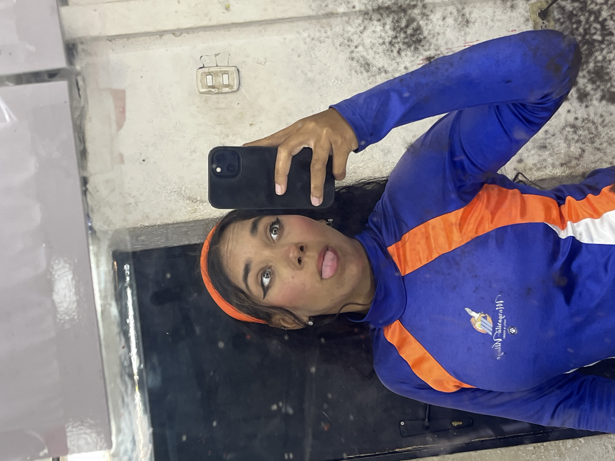 A woman with dark hair and olive skin wearing a blue and orange long-sleeved shirt and a orange headband is taking a selfie in what appears to be a dirty mirror, she's sticking her tongue out. The shirt has a logo on it that appears to read "Margarita Viva" with the words "Nueva Esparta" underneath. This logo indicates that the location is Porlamar, Venezuela, as Margarita is a popular vacation destination in Nueva Esparta, Venezuela. Behind her, one can see an electrical outlet on the wall and a possible interior.