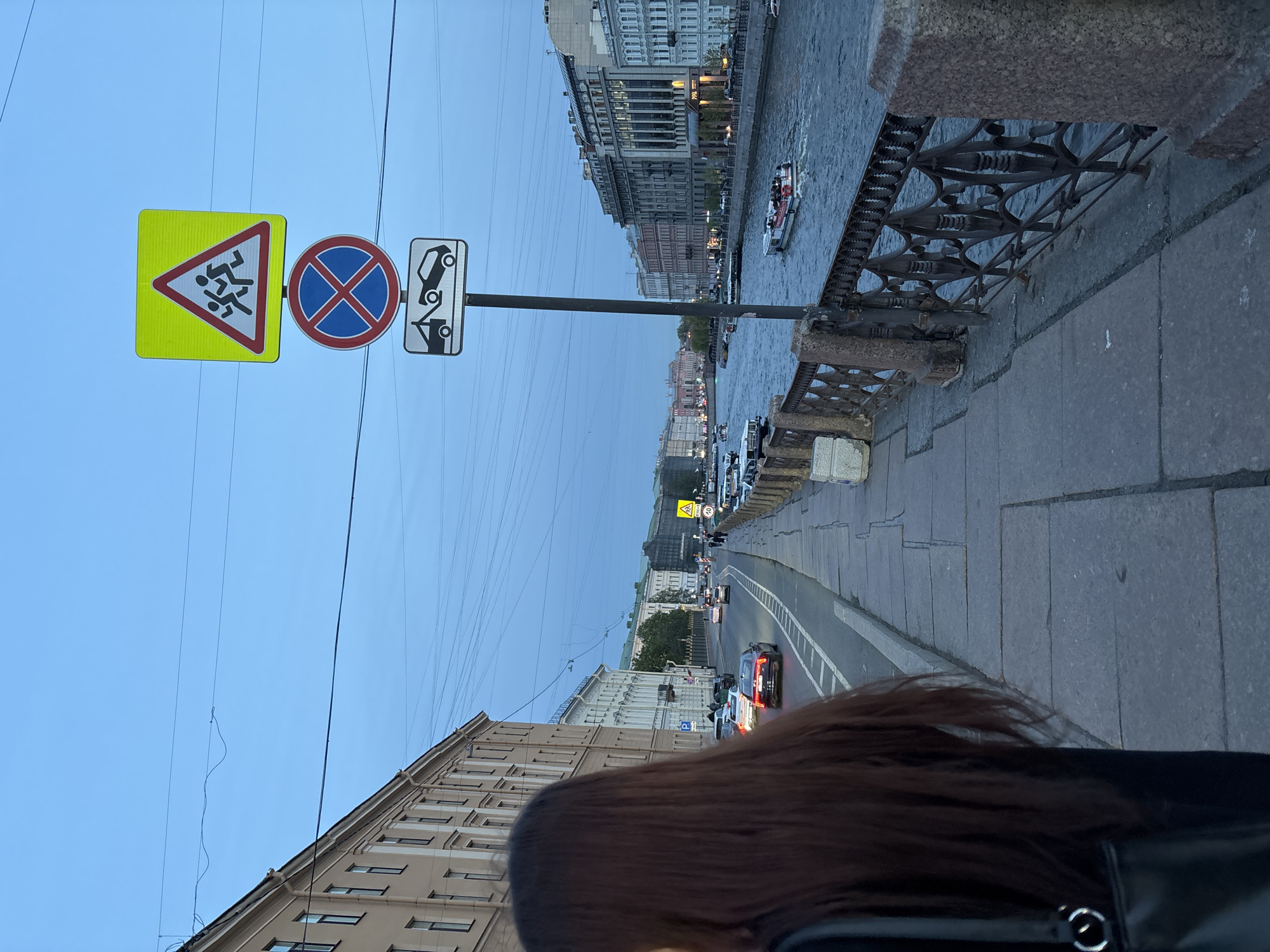 The image depicts a street scene in Saint Petersburg, Russia, taken from an elevated position, likely a bridge overlooking a waterway. The camera angle is oblique.
The scene includes traffic and pedestrian activity along a city street, and boats on the river.
In the foreground, a woman's head and hair is visible. To the left are multi-story buildings.
A signpost stands on the left, displaying three traffic signs, including a pedestrian crossing warning sign, a 'no stopping' sign and a 'no parking' sign.
Beyond the street, a river flows through the city, with buildings lining its banks. Several boats are on the water. The overall lighting suggests it is either early morning or late evening.