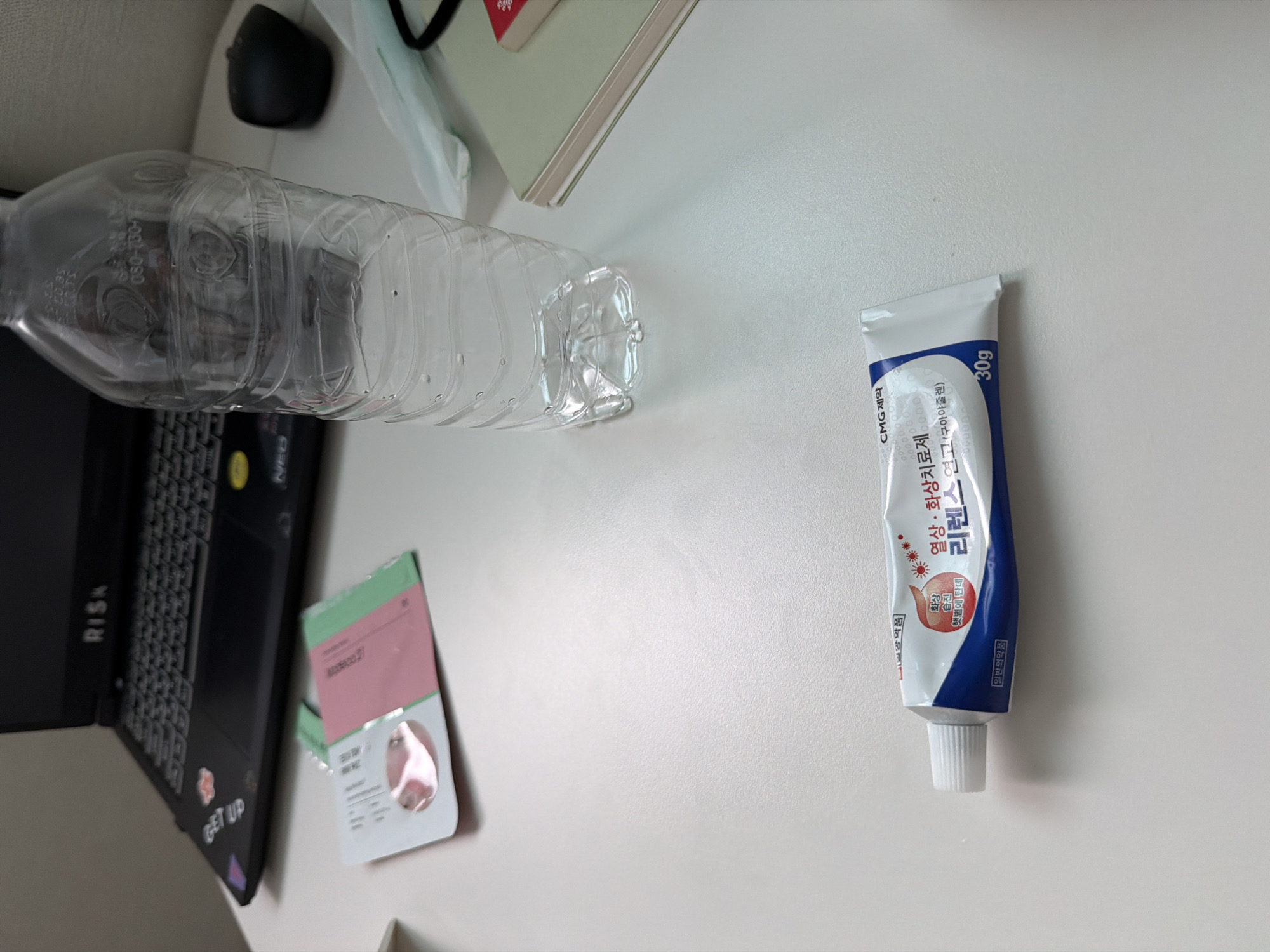 The image shows a desk with a laptop, a water bottle, and a tube of ointment. The laptop is black and has stickers on it. The water bottle is clear and empty. The ointment tube is white and blue and has Korean text on it. The scene appears to be indoors, possibly in an office or home. There is no visible indication of the city of Dongducheon-si, South Korea.