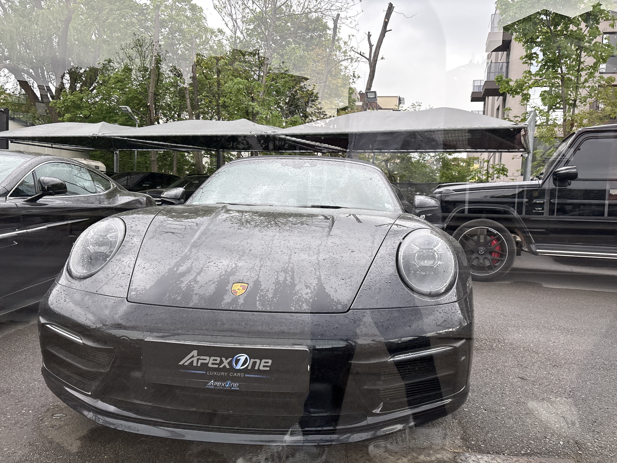 A rainy day in Sofia, Bulgaria shows three luxury cars under a parking awning. The closest car is a black Porsche 911 with the license plate partially visible showing "Apex One Luxury Cars." Behind the Porsche is a dark gray or black coupe. To the right of the Porsche is a black Mercedes-Benz G-Class SUV with red brake calipers. In the background are trees and a building with balconies.