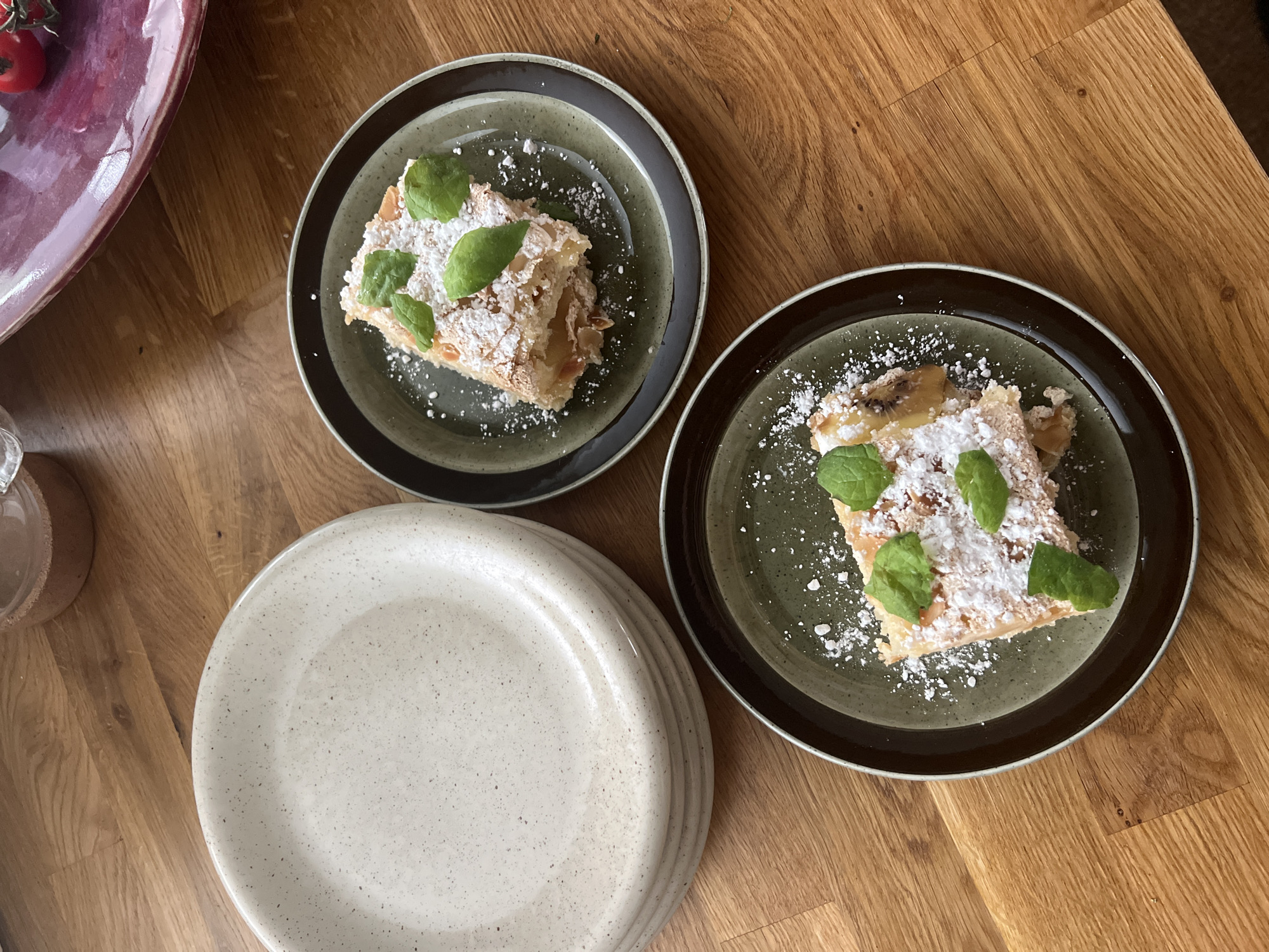 A stack of speckled, light-colored plates sits on a wooden table. To the right of the plates, two dark-rimmed, green plates each hold a slice of strudel, dusted with powdered sugar and garnished with fresh mint leaves. The strudel appears to have a flaky pastry crust and a filling of sliced fruit. The overall scene suggests a dessert being served for enjoyment. This image was taken in Hämeenlinna, Finland.