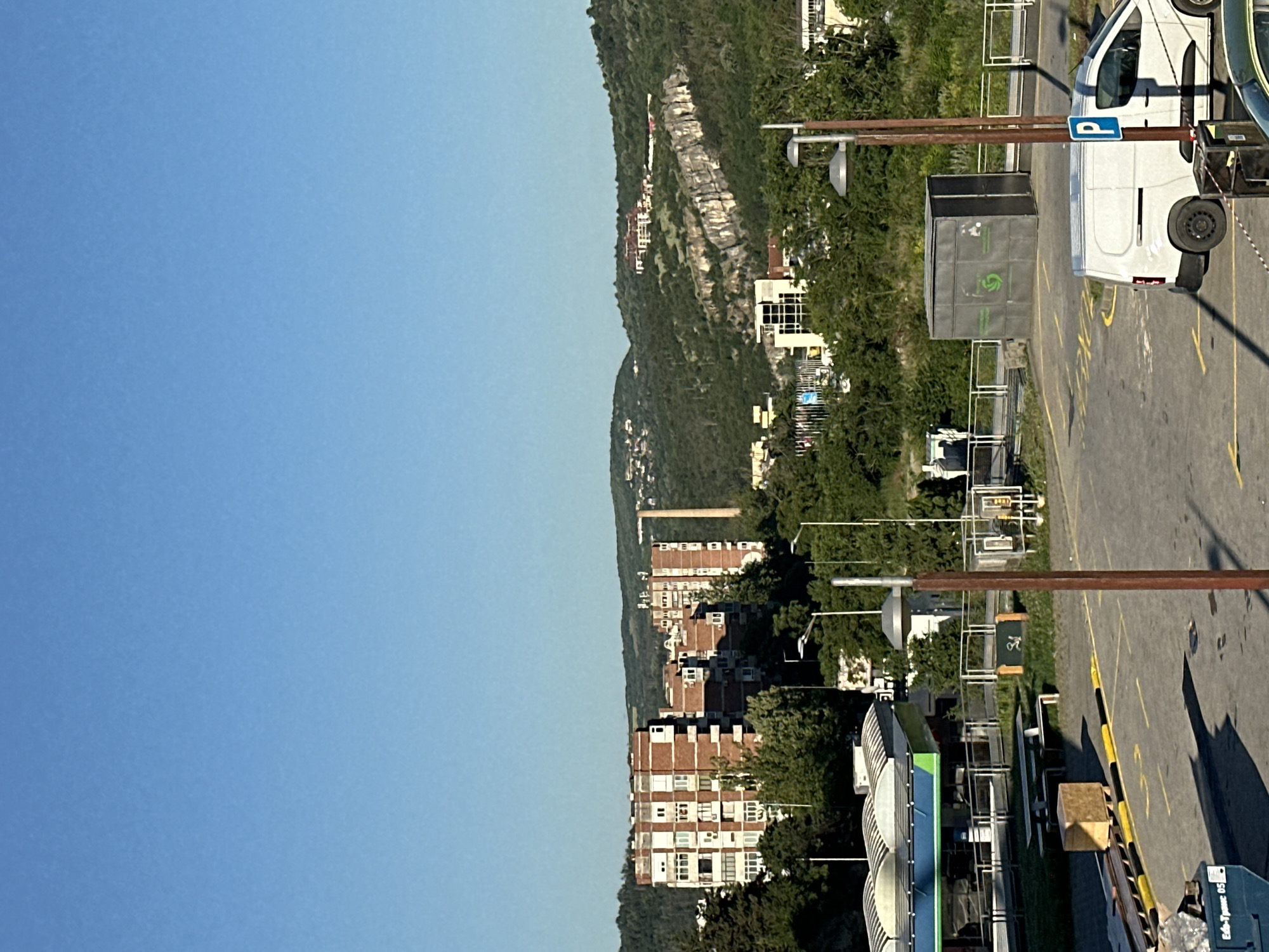 The image shows an outdoor urban scene under a clear blue sky. In the foreground, a paved parking lot is visible with yellow markings. A white van is parked on the right side of the lot, facing left. A blue parking sign with a white 'P' is mounted on a pole next to the van. To the left, a blue dumpster is visible. Further back, a structure resembling a small industrial building or storage unit with green symbols on its side is situated near a metal fence. Several streetlights with curved shades are also present. Beyond the parking lot, a gas station with a curved roof and blue and green accents is visible. A picnic table and a wooden crate are placed on a grassy area near the gas station. In the mid-ground, lush green trees dominate the landscape, interspersed with various buildings. Among the trees, several apartment buildings are prominent, characterized by red brick exteriors and numerous windows. A tall, slender smokestack or industrial chimney stands behind some of these buildings. In the background, rolling hills covered in dense vegetation form the horizon. Several buildings are scattered on the slopes of these hills, with some appearing to be on the crest. A distinct rock formation with sheer, light-colored cliffs is visible on one of the hillsides. The overall impression is a sunlit day in an area that combines residential, commercial, and natural elements. Based on the architecture of the buildings, particularly the apartment blocks, and the natural landscape with hills and cliffs, this scene is likely located in the city of Veliko Tarnovo, Bulgaria, a city known for its hilly terrain and historical architecture.