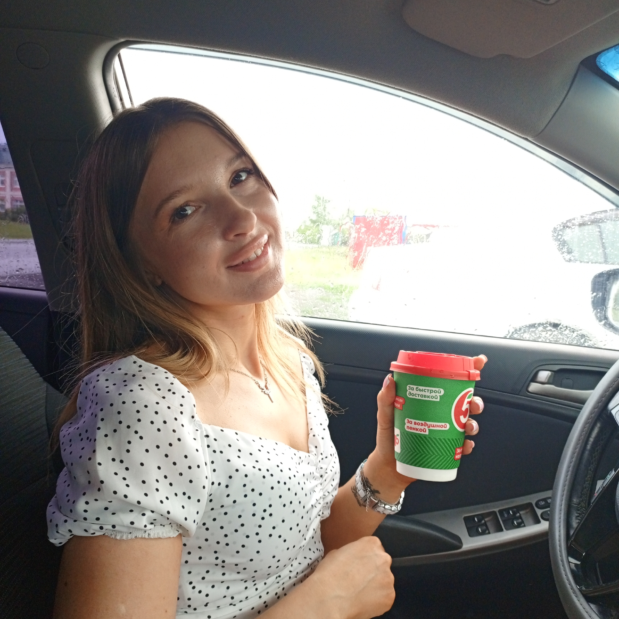 A young Caucasian woman with blonde hair and a polka-dot dress is seated in the driver's seat of a car, holding a green and red disposable coffee cup. The cup has Russian text that translates to "For fast delivery" and "For airy foam." The car's interior is visible, including the steering wheel, door panel with window controls, and a side mirror. Outside the car window, a blurry cityscape with buildings and trees can be seen, with raindrops on the glass indicating rain. The scene is set in Petukhovo, Russia.