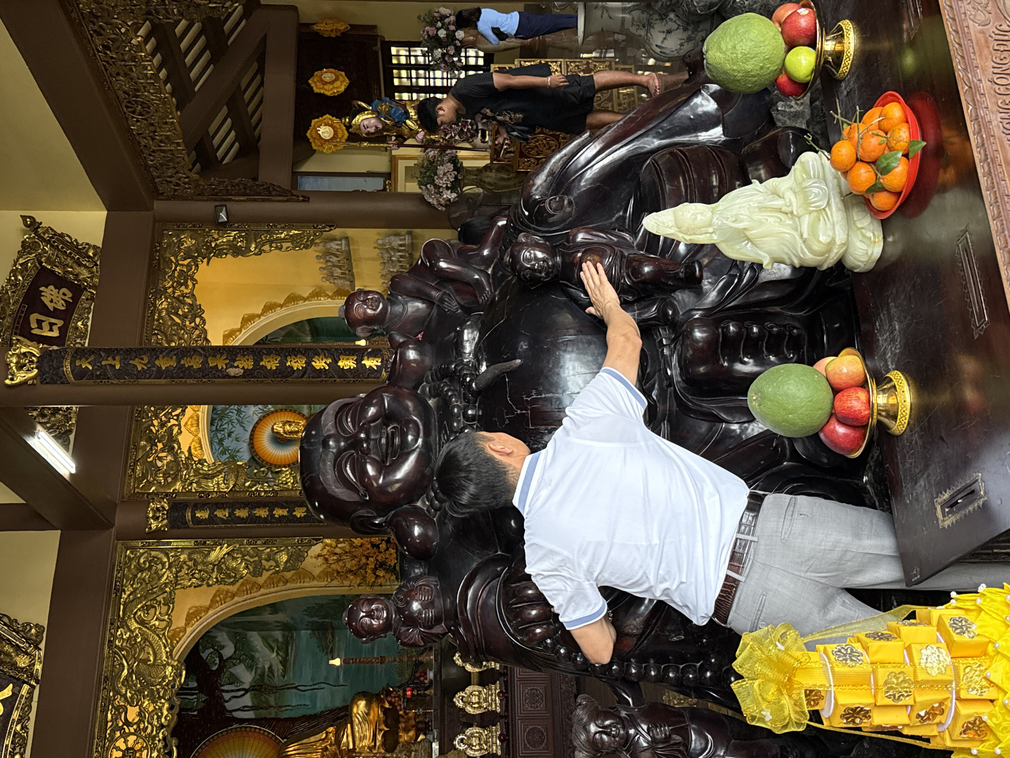 The image depicts the interior of a Buddhist temple, identified as Phap Lam Pagoda, located in Da Nang, Vietnam. The central focus is a large, dark-colored, possibly wooden or lacquered, statue of a corpulent, laughing Maitreya Buddha, surrounded by several smaller dark figures.
In the foreground, a dark wooden altar table displays various offerings. On the left, a tall, multi-tiered yellow gift package with a large bow is visible. Two large green pomelos or grapefruits, one on the left (partially obscured) and one on the right, rest on golden pedestals, accompanied by red apples and a small green lime. A translucent, light-colored statue of a seated Guanyin Bodhisattva is centrally placed on the table, and a red bowl filled with tangerines, some with leaves, is in the lower right.
A middle-aged Asian male, dressed in a white polo shirt with light blue trim and light grey trousers, stands in the lower-middle left, facing away from the camera. He is leaning slightly forward with both hands placed on the abdomen of the large Maitreya Buddha statue. In the mid-ground right, a younger Asian male with dark hair, wearing a black t-shirt and black shorts, stands barefoot. He holds a white rectangular object, possibly a mobile phone, in his left hand. Further in the background right, a woman in a light blue shirt and dark pants is partially visible, facing a window.
The temple interior is elaborately decorated with extensive gilded carvings on pillars, archways, and wall panels, depicting intricate patterns, including dragons and floral motifs. Sections of murals depicting landscapes are visible through gilded archways. Gilded calligraphy is present on columns and overhead beams, including "佛日" (Fó Rì). Various other statues are scattered throughout the background, including smaller golden Buddhas and a standing golden figure with blue accents. Artificial floral arrangements in large vases are also visible. The ceiling features dark wooden beams and a modern fluorescent light fixture.