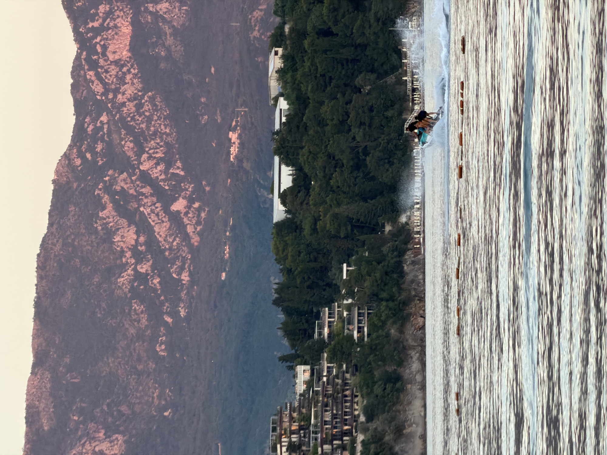 The image shows a scene in Budva, Montenegro, with a mountain range in the background and a body of water in the foreground. Along the shoreline, there are residential buildings nestled amongst trees. In the water, a small group of people are riding a wakeboard being pulled by a motorboat. The water is lined with buoys, likely marking a designated area for water sports.