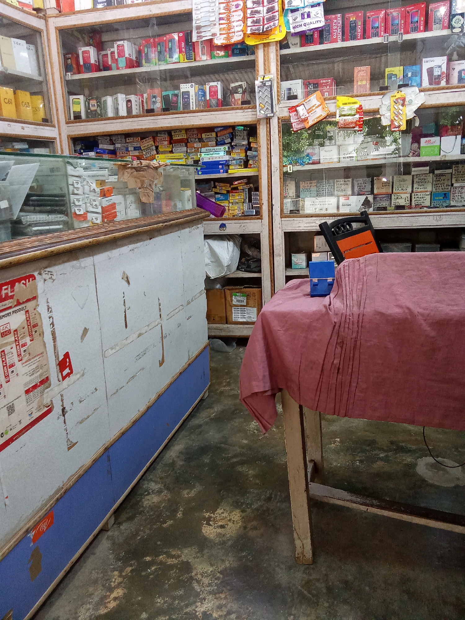The image depicts a small retail shop, likely an electronics store, in Konta, India. The shop is filled with shelves packed with various electronic items, including mobile phones, accessories, and possibly other small electronic devices. There are clear plastic display cases showcasing some of the merchandise. On the left side of the image, a counter is visible with some stickers on its surface, and a glass display case on top. The flooring appears to be concrete with some discoloration. A table covered with a pink cloth is positioned in the foreground, obscuring some of the shop's contents. On this table, a black and orange electronic device and a small blue object are placed. The overall impression is of a busy, somewhat cluttered, and well-stocked local business.