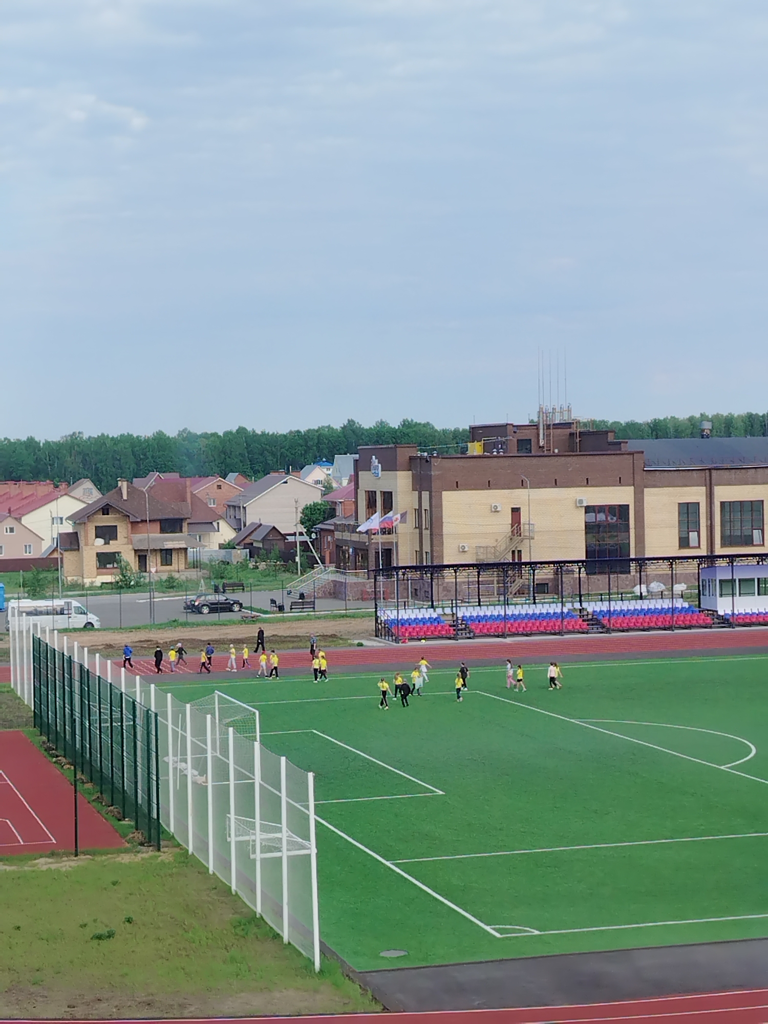 The image shows a soccer field and running track in Noviy, Russia. The field is green and has white lines marking the playing area. People are present on the field, some wearing yellow shirts. A track surrounds the field, with red and grey surfaces. A building is visible in the background, with an outdoor staircase. There are also several houses with red roofs, as well as green trees.