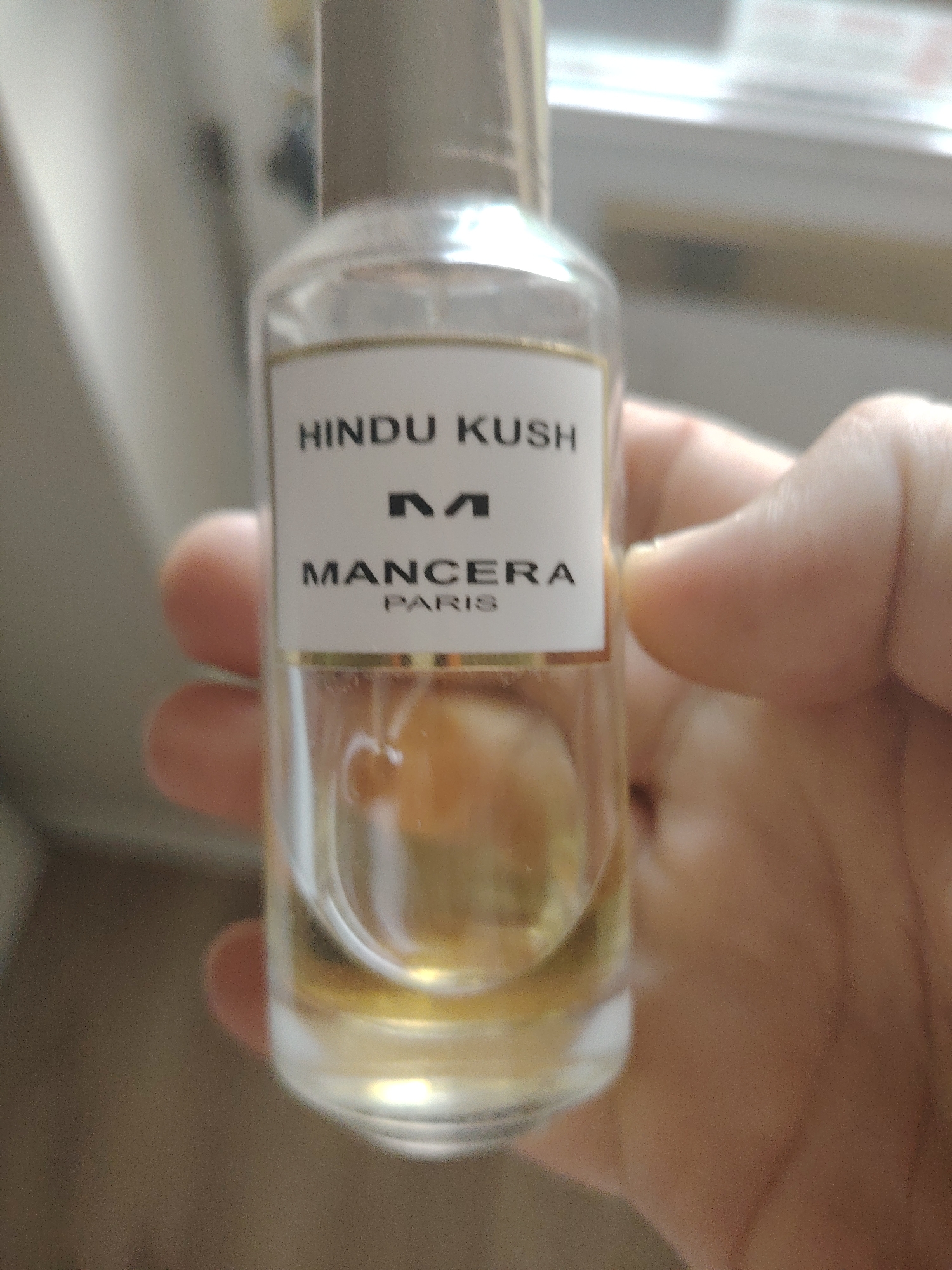 A partially obscured, clear glass perfume bottle with a metallic cap is held in a person's hand. The bottle contains a golden-colored liquid. A white label on the bottle displays the text "HINDU KUSH", a logo with an "M", and then "MANCERA" followed by "PARIS". The background is blurred and appears to be indoors, possibly near a window. There is no discernible location in Huntingdonshire, United Kingdom.