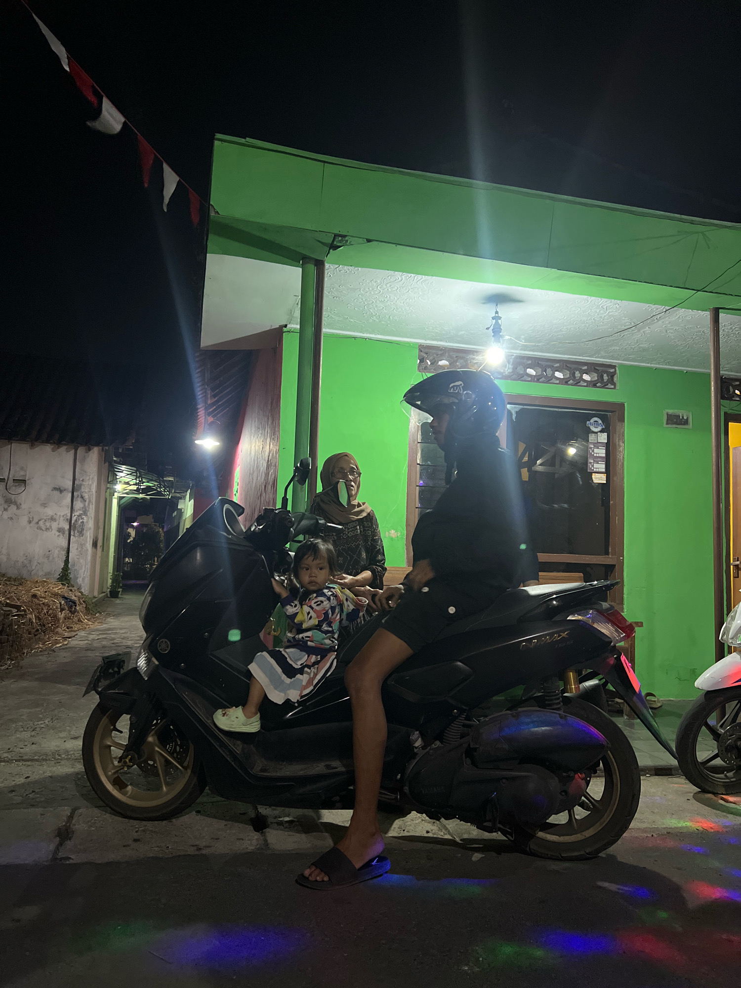 A family of three is on a dark-colored scooter at night. A person in a helmet and black clothing is driving. A child in a colorful outfit is sitting between the driver and a woman wearing a headscarf and glasses. They are parked on a street in front of a building with green walls and a dark roof. Red and white flags are strung across the top left of the image. Colorful lights are reflected on the ground in front of the scooter.