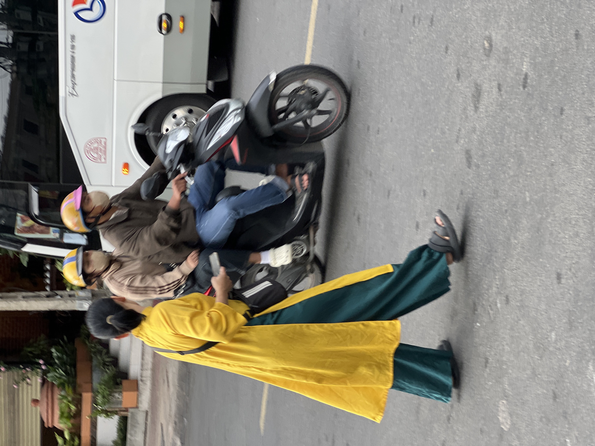 The image captures a street scene in Nha Trang, Vietnam, likely near a bus terminal or a main thoroughfare given the presence of a large white bus. In the foreground, a woman walks away from the camera, wearing a bright yellow, floor-length tunic over dark green wide-leg pants. She has her dark hair tied back and carries a black crossbody bag with white stripes. Her feet are clad in dark sandals. Behind her, two individuals are riding a dark-colored motorcycle. The person in front is driving and wears a brown jacket, blue jeans, and a colorful helmet. The person behind, a passenger, wears a beige jacket, a yellow and blue helmet, and a face mask. The motorcycle has red accents and is in motion. The bus in the background is white with "Empress 1615" printed on its side and features a logo with blue and red elements. The road surface appears to be asphalt, with some faint markings visible. The background shows some greenery and architectural elements, suggesting an urban environment.