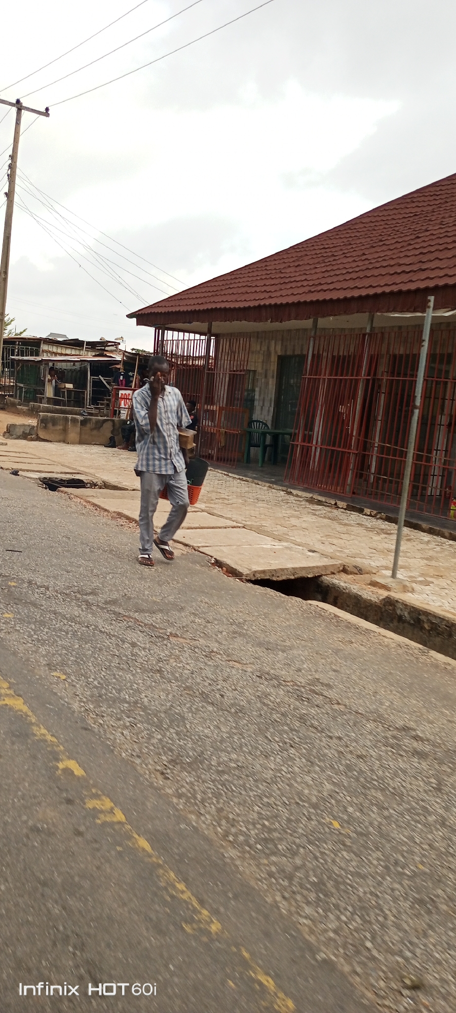 The image shows a man walking on a street in Adankolo, Nigeria. He is wearing a blue and white checkered shirt and grey trousers, and carrying items in his hands. The street has yellow lines and a damaged concrete walkway with exposed drainage underneath. Behind the man is a building with a brown roof and red bars on the windows. In the background, there is a street vendor structure. Overhead, there are utility lines. The sky is overcast.