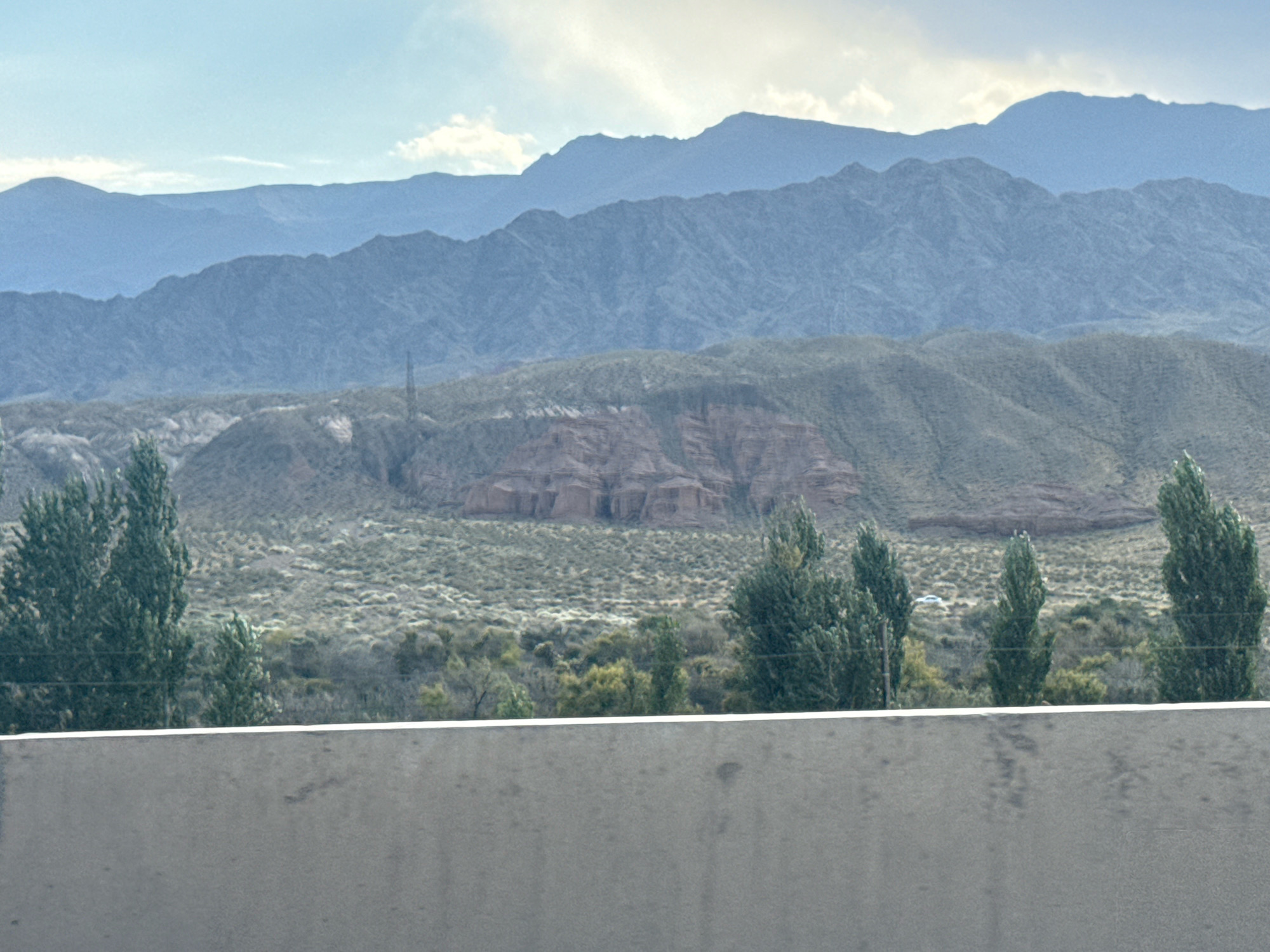 The image depicts a distant, mountainous landscape with layers of receding mountain ranges under a pale sky. In the mid-ground, a semi-arid terrain is visible, characterized by scrubby vegetation and rock formations. Several tall, slender trees are scattered across the foreground and mid-ground, their dark green foliage contrasting with the muted earth tones of the landscape. In the immediate foreground, a concrete barrier runs horizontally across the frame, partially obscuring the view of the natural scenery. The overall impression is one of a vast, undeveloped natural environment, likely a rural or remote area.