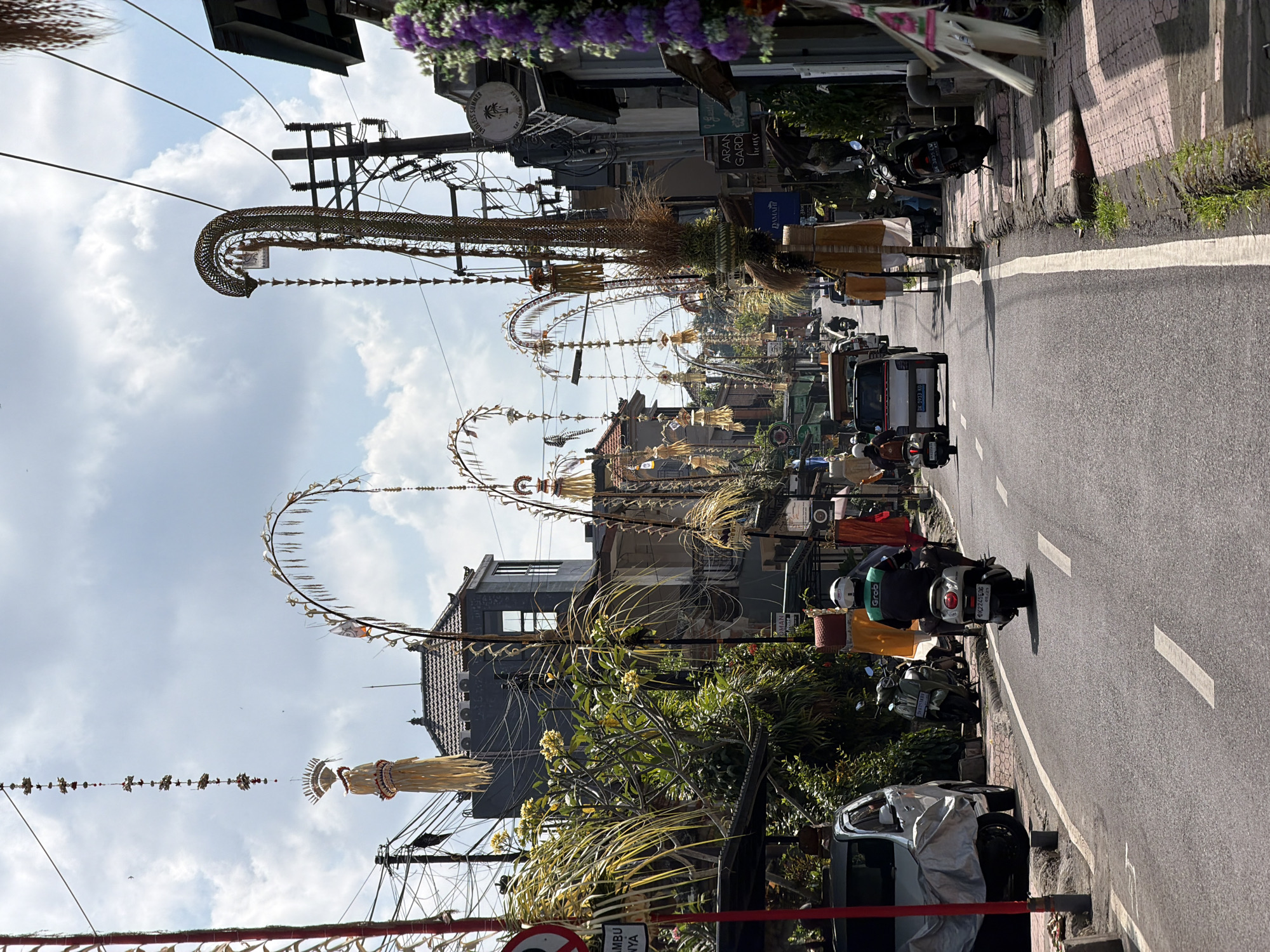 This image captures a street scene in Ubud District, Indonesia, characterized by a procession of decorative banners and arches hanging overhead, adorning the roadway. A silver van is parked on the left, partially covered with a tarp, and a scooter with a rider wearing a white helmet and a green jacket, branded with "Grab", is proceeding down the asphalt road. Several other scooters and a white vehicle are visible further down the street. The road is lined with buildings, some of which appear to be shops or residences, and lush green vegetation. The sky above is partly cloudy, suggesting a bright, sunny day. The overall impression is of a vibrant street in a bustling town, possibly during a local festival or celebration, given the abundant decorations.