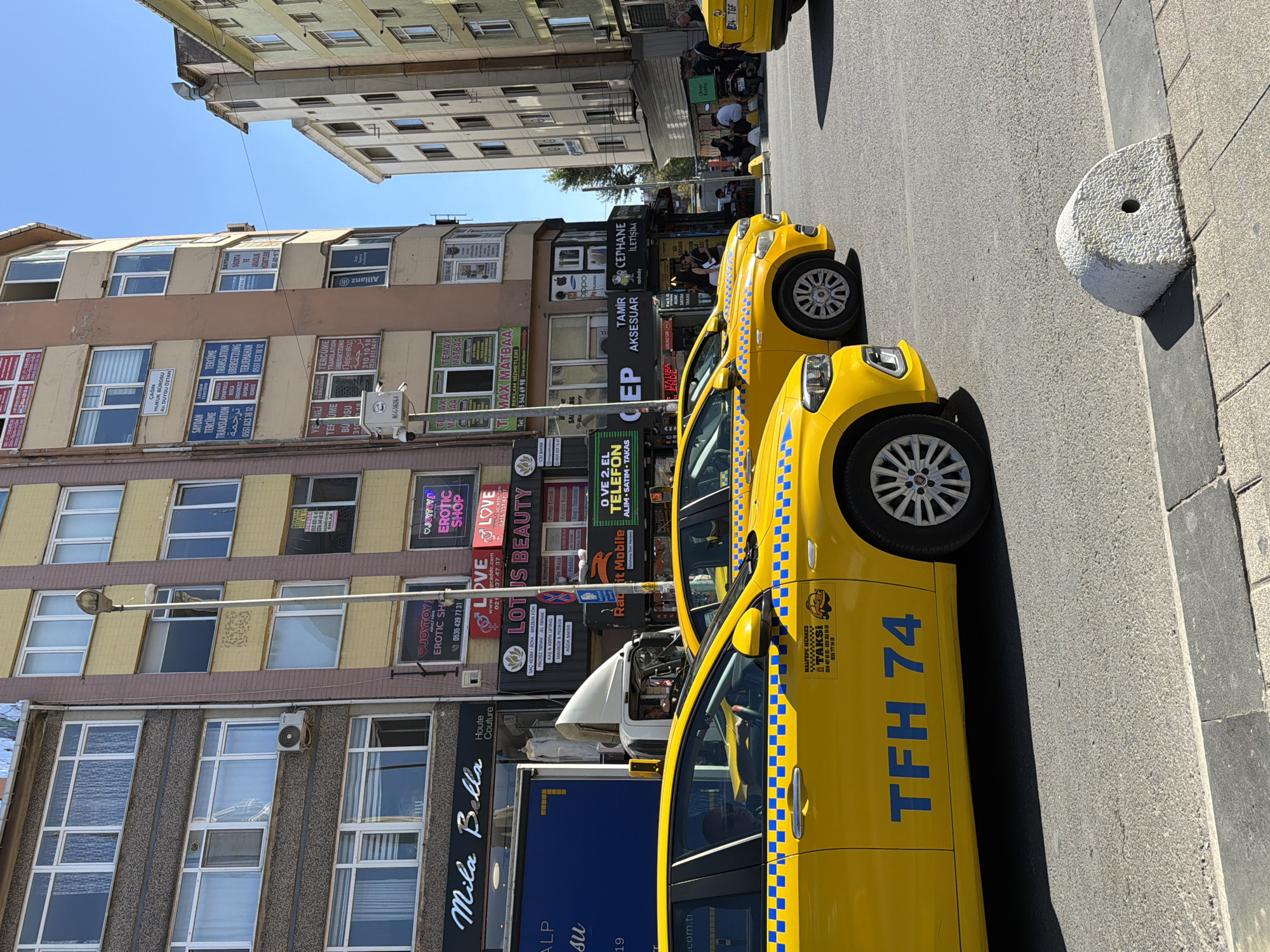 The image depicts a street scene in Kadıköy, Turkey, under a clear blue sky. In the foreground, two bright yellow taxis are parked on the asphalt road. The taxi on the left has "TFH 74" written in blue on its side. A white, dome-shaped concrete bollard with a circular hole is visible on the sidewalk in front of the taxis.
Behind the taxis, a multi-story building with a beige and brown facade dominates the background. Various signs and advertisements are affixed to the building's lower levels. Notably, there's a sign for "Mila Bella Haute Couture." Other signs include "EROTIC SHOP," "JOYTOY," and "LOTUS BEAUTY." A white truck with a visible driver is parked behind the first taxi. To the right of the main building, a taller, lighter-colored building is partially visible, along with a sliver of an even taller, pale building further to the right. A few people are gathered on the sidewalk in front of the buildings, and a scooter with a delivery box is parked nearby. The overall impression is of a busy urban commercial area.