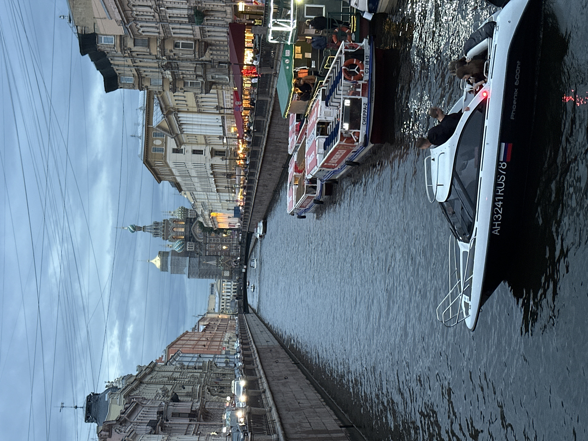 This is an exterior, eye-level, medium shot photograph of a canal with boats and buildings in St. Petersburg, Russia. The sky is overcast and the lighting is dim, suggesting it is either early morning or evening.
In the foreground, a white speed boat with a black hull is moving from right to left, carrying four people. Two men are seated in the front of the boat, while two women are seated in the back. The boat has the registration number AH3241RUS78 on its side.
Further down the canal, several tour boats are docked. These boats are red and white, with "СИЕРРА-2" written on their sides. They appear to be carrying passengers.
The canal is lined with stone walls. On the left side of the canal, there are multi-story buildings with a variety of architectural styles. On the right side, there are more buildings, some of which have awnings and illuminated windows, suggesting they are shops or restaurants.
In the background, a large, ornate cathedral with a green dome and multiple spires can be seen. This is likely the Church of the Savior on Spilled Blood, a prominent landmark in St. Petersburg. Overhead, numerous electrical wires are visible against the cloudy sky. Traffic can be seen on the street running alongside the canal on the left.