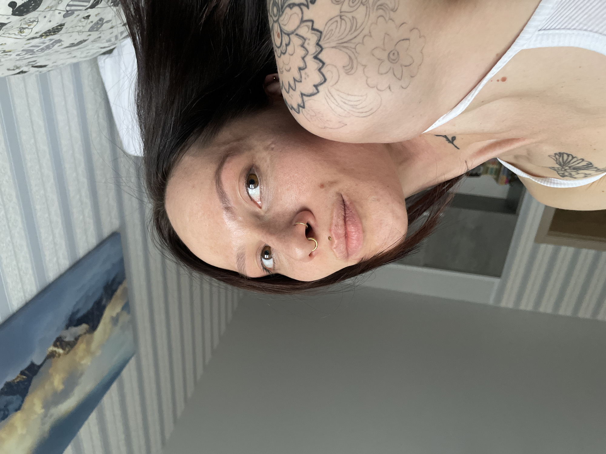 The scene depicts a female individual in an indoor, likely residential, setting located in Izhevsk, Russia. The subject, an adult female with long, dark brown hair, is positioned diagonally, facing upwards towards the camera. She is wearing a white, ribbed tank top or camisole.
Visible on her body are multiple tattoos and piercings. A dark silhouette tattoo of a bird, possibly a hummingbird, is on her right collarbone area. On her left shoulder/upper arm, an unshaded, outlined floral or mandala-like design is present. A small, dark tattoo fragment is also visible on the right side of her neck/upper back. Her face features a gold-colored hoop piercing in her right nostril and a small gold stud piercing above her upper lip. Her brown eyes are directed towards the camera. A reddish mark is visible on her right collarbone.
The immediate environment consists of a room with light-colored wallpaper featuring vertical grey and white stripes. The ceiling is plain light grey. To the right of the subject, a white pillow and a duvet or blanket with a light background and dark patterns of animals or characters are visible, suggesting a bed. A rectangular painting depicting a mountain or cloudscape in blues, whites, and yellows/golds is hung on the striped wall to the right. On the far left, a door frame with indistinct shapes beyond it is partially visible. The lighting is even, suggesting ambient or natural light.