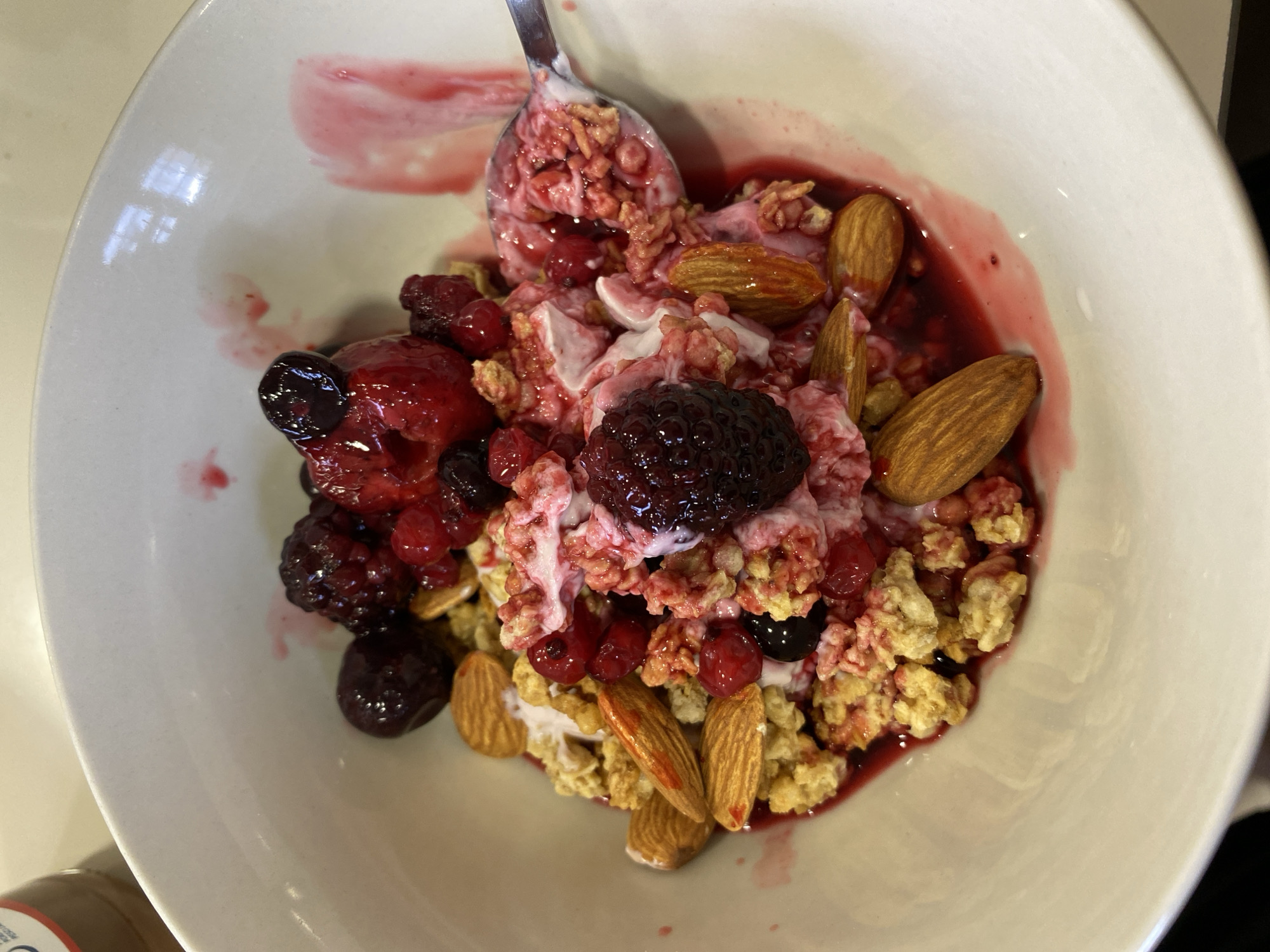 The image depicts a white bowl filled with a breakfast or snack meal. The meal consists of granola, several whole almonds, mixed berries (including blackberries, cherries and cranberries), and yogurt. A spoon, also coated with the meal, is inside the bowl, suggesting it has recently been eaten. There is no clear indication the image was taken in Villaviciosa, Spain, or any other specific location. The environment is unspecified, but the bowl rests on a light-colored surface. There are no people visible.
