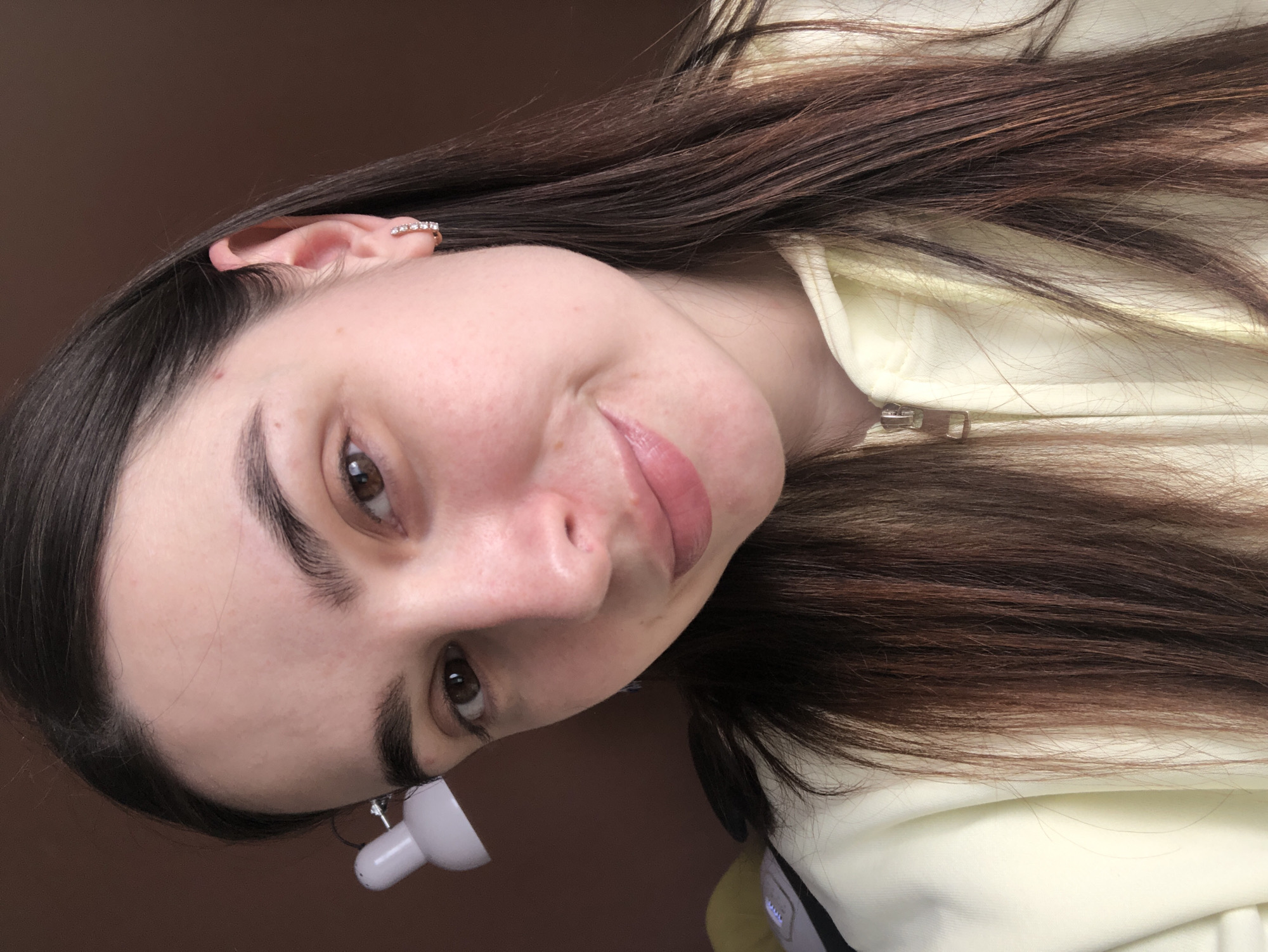 A close-up portrait captures a young woman, likely in a selfie. She has long, straight dark brown hair, parted around the middle, flowing over her shoulders and a light yellow garment. Her head is tilted to her right, and she looks directly at the camera with a gentle, subtle smile, conveying a calm and friendly expression. She has dark brown eyes and well-defined eyebrows. Her lips are a natural pink hue, slightly parted.
She wears a light yellow top, possibly a hoodie or jacket, with a silver-toned zipper pull visible. On her visible ear, she sports a delicate ear cuff adorned with small, clear, sparkling stones. A small, white, cylindrical object with a darker tip, resembling a vape pen or similar personal electronic device, is partially visible near her chin and neck.
The background is a plain, solid warm brown color, indicating an indoor setting with a simple wall or furniture. The lighting is soft and even, without harsh shadows. No text is visible in the image. Although the photo lacks specific geographical cues, it was captured in Chaikovsky, Russia.