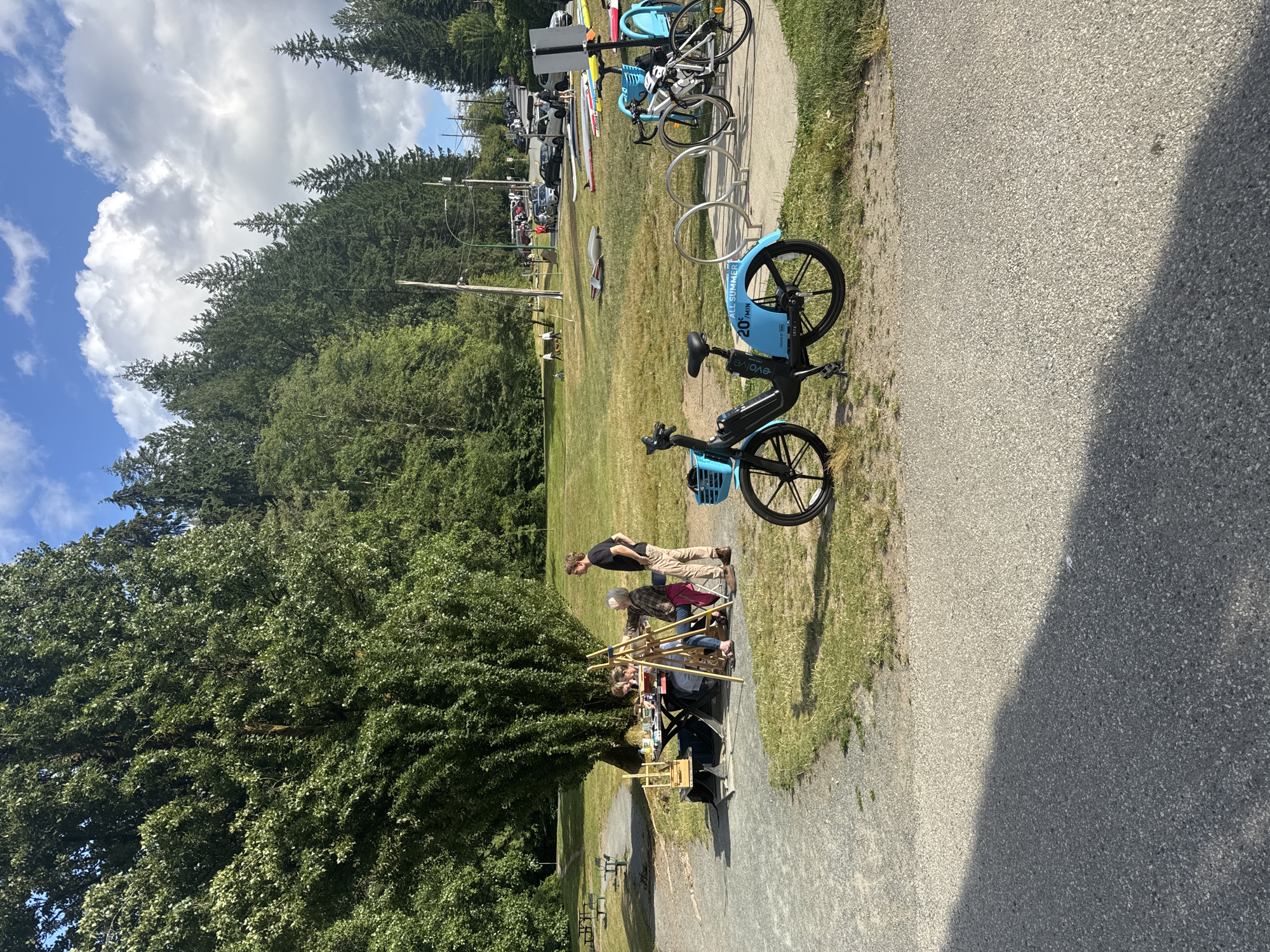 A sunny daytime outdoor scene is captured at Mosquito Creek Park in the District of North Vancouver, Canada. A group of people are gathered around a picnic table under a large, mature tree, engaged in what appears to be an art session, with easels and art supplies visible. A man in a black t-shirt and tan pants stands near the group, observing. In the foreground, a blue shared bicycle is parked, with a bike rack holding two other bicycles in the mid-ground. Beyond the park area, a street with parked cars and people walking is visible, with a densely wooded hillside and a cloudy sky forming the backdrop. The ground is a mix of gravel paths and grassy areas.