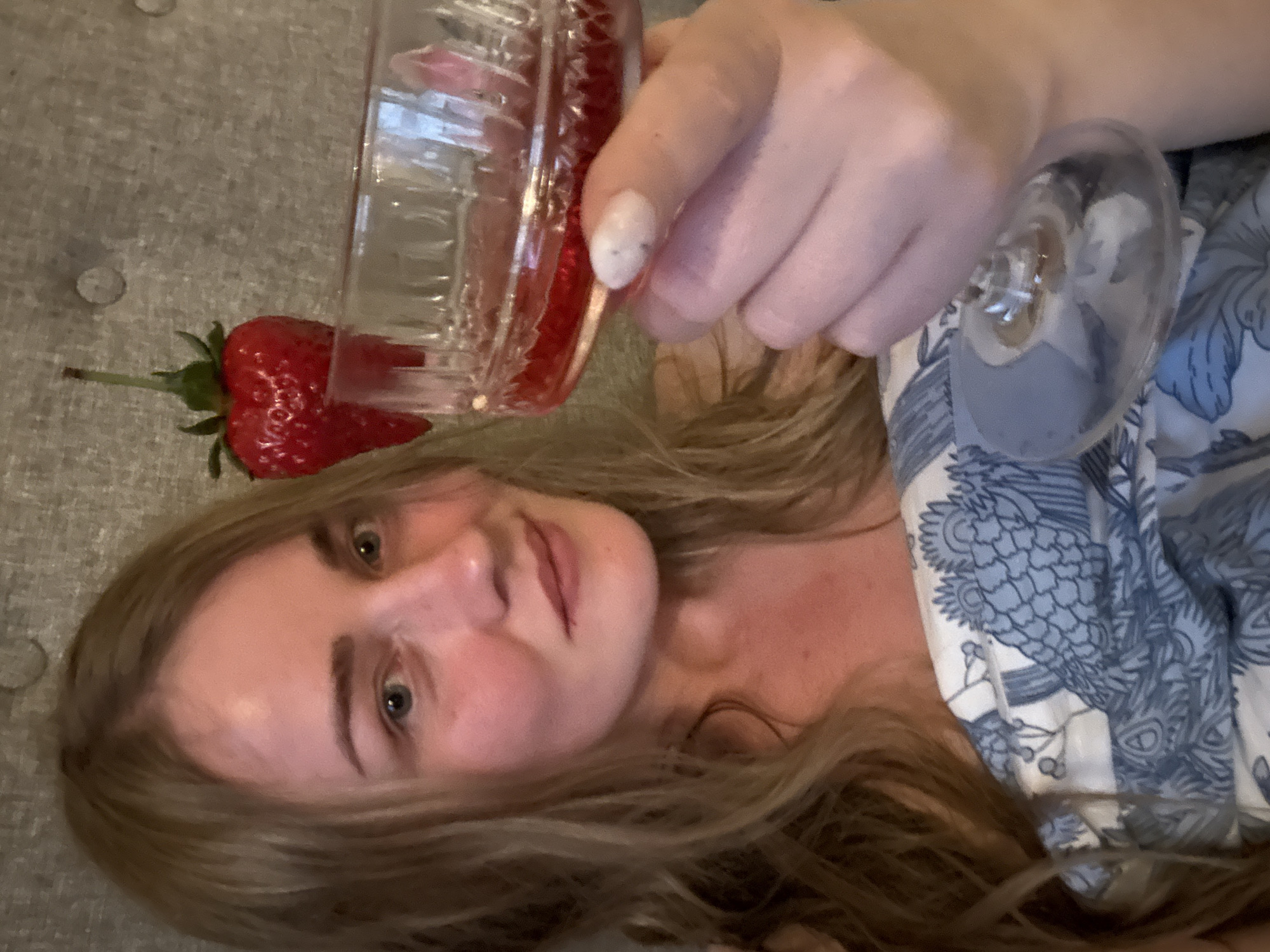 A woman with long blonde hair is lying on a gray button-tufted surface. She is holding a crystal glass filled with a red liquid. A large strawberry with green leaves is next to the glass on the left. She is wearing a blue and white patterned garment.